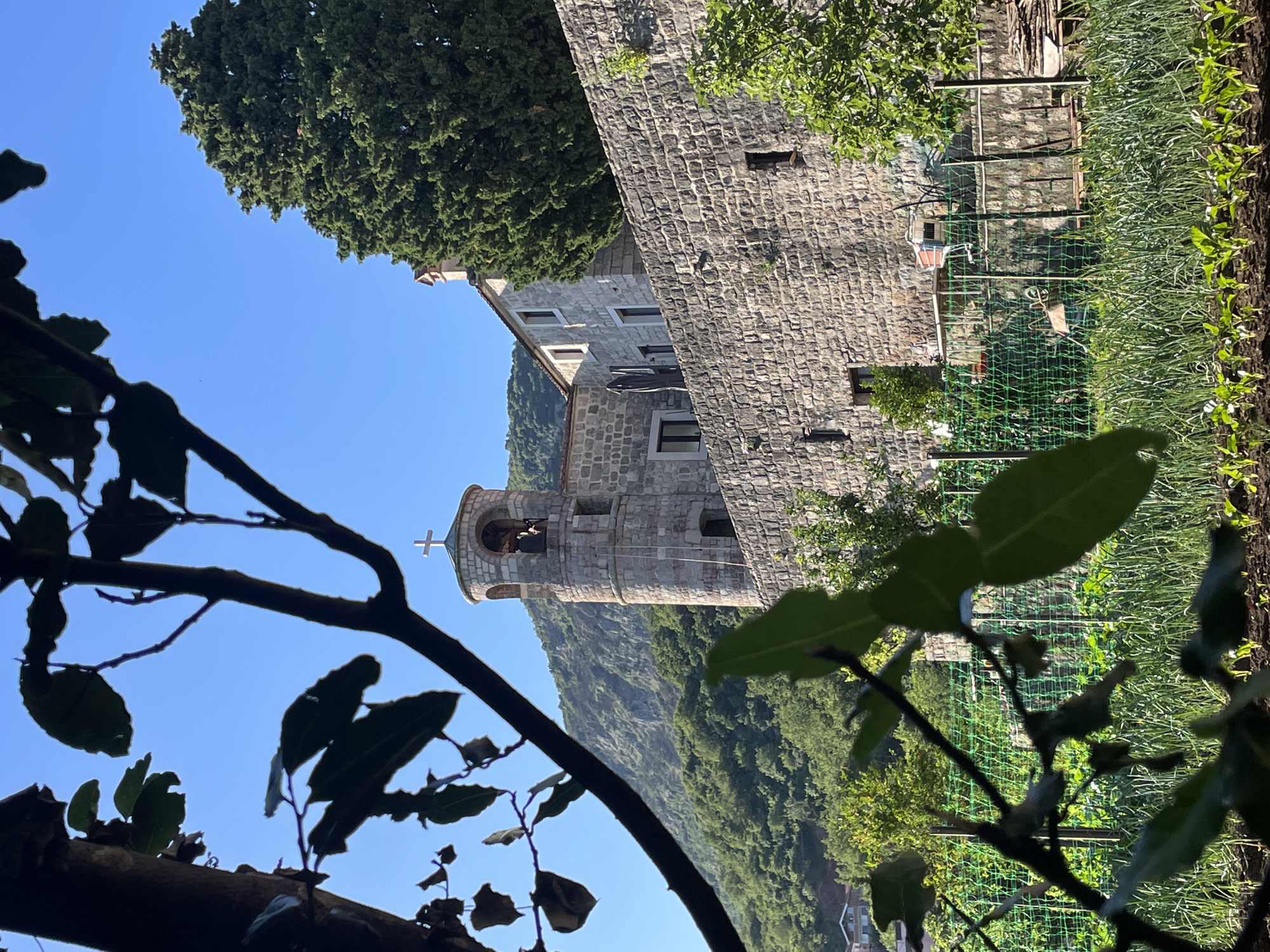 The image shows the Praskvica Monastery in Budva, Montenegro, viewed from a low angle through foliage. The monastery's stone buildings rise against a bright blue sky, with a prominent bell tower topped by a cross. The surrounding landscape includes a lush green hillside and what appears to be a garden or cultivated area with fencing. The foreground is dominated by the dark silhouettes of leaves and branches, adding depth to the composition.