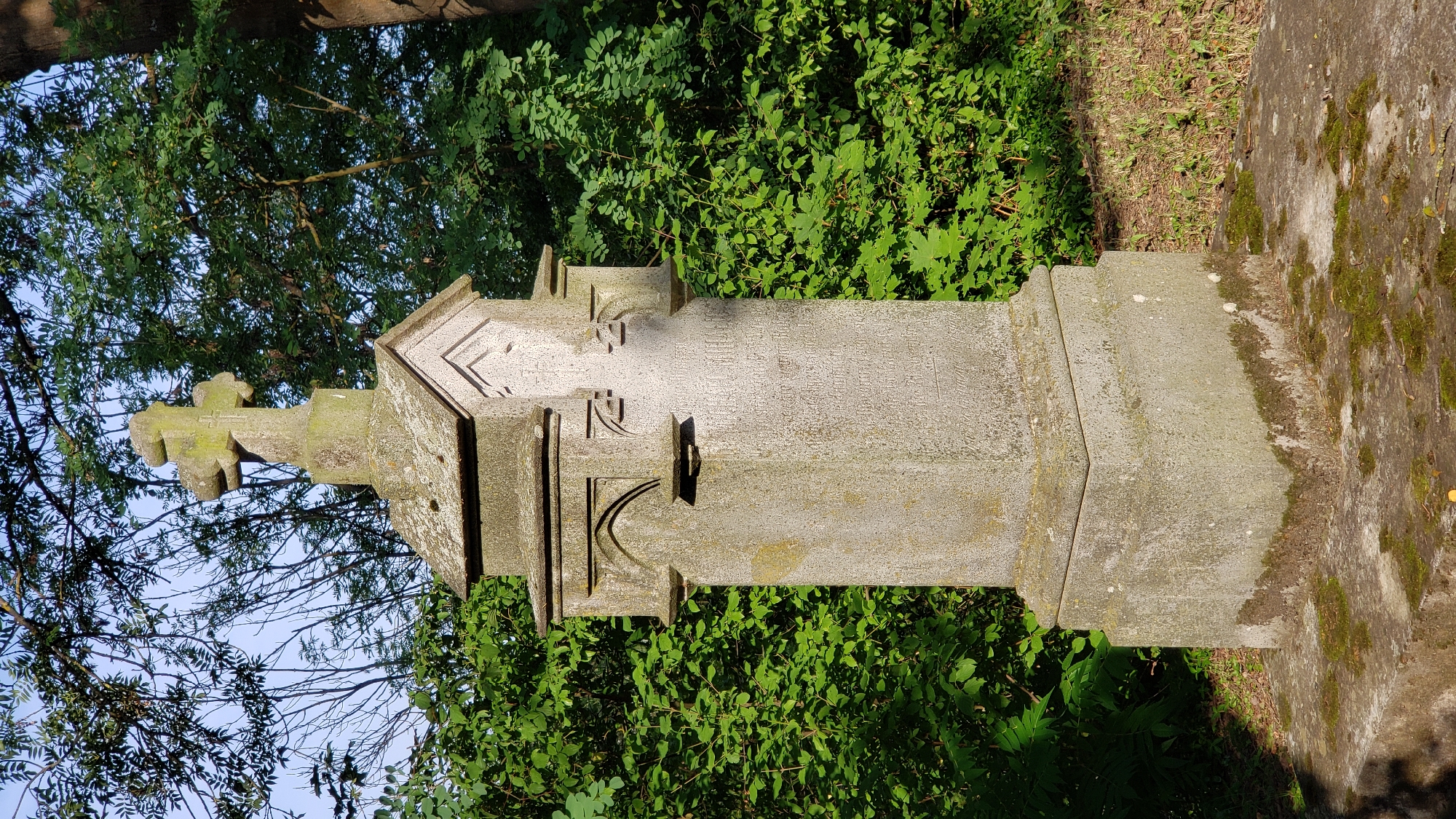 The image shows a weathered stone cross atop a tombstone in a green, wooded area, likely a cemetery. The tombstone is elaborately carved with architectural details, including a roof-like structure at the top and decorative arches. Inscriptions are visible on the main body of the tombstone, though they are difficult to read due to age and wear. The cross itself also shows signs of weathering and moss growth. The surrounding environment consists of lush green foliage and trees, suggesting it is a natural or park-like setting. The lighting indicates it is daytime, with sunlight filtering through the leaves. The scene appears peaceful and solemn. This specific tombstone is located in the Catholic Cemetery (Katolícky cintorín) in Prešov, Slovakia, and is a memorial to a person named "STEFAN KORPACSY".