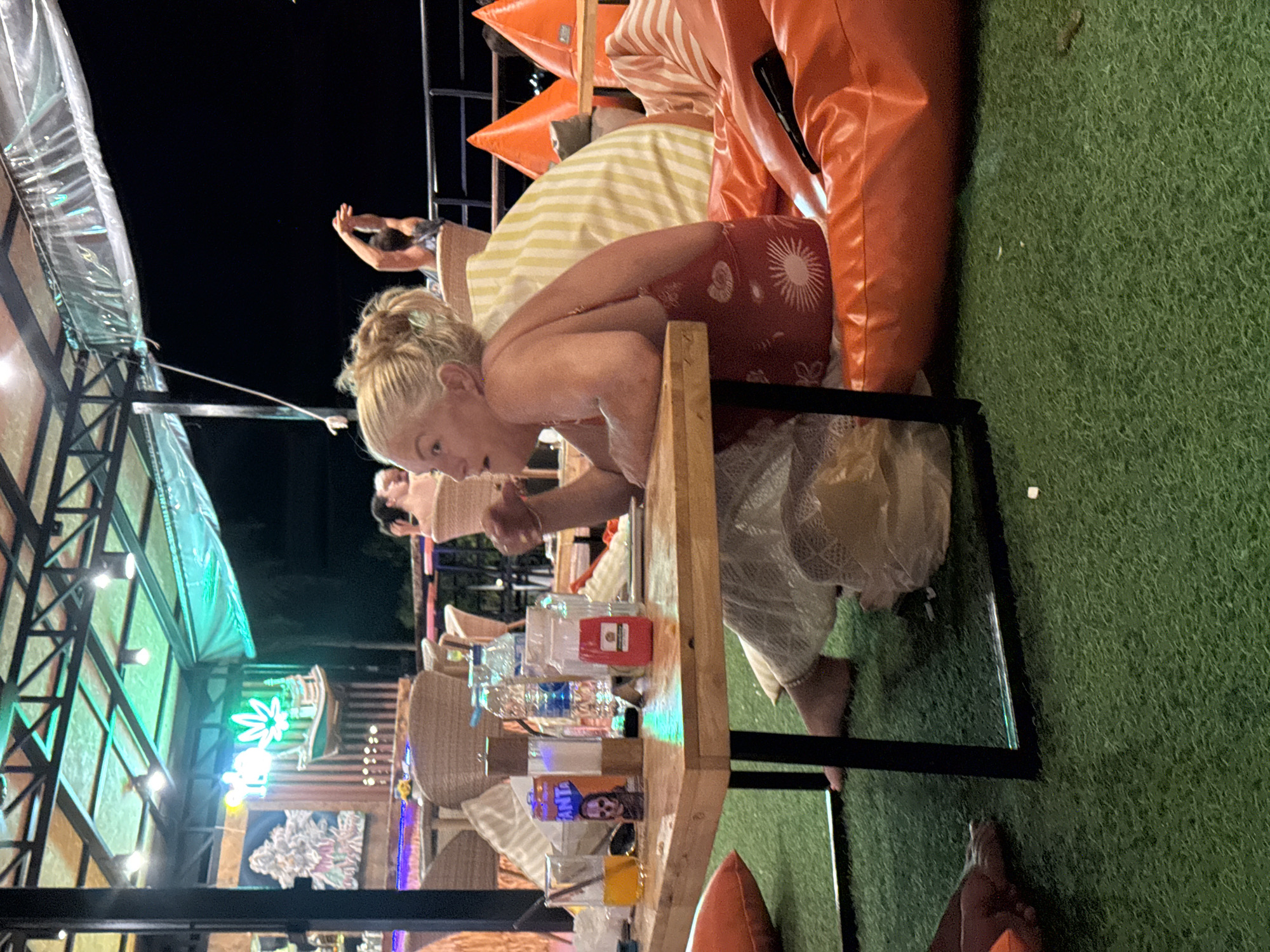 The image captures a nighttime scene at an outdoor establishment in Ko Samui District, Thailand. A blonde woman in a red dress is seated at a wooden table, looking to her left. On the table are drinks, including a glass of orange liquid and bottles of water, as well as a can with an image of a skull. She is sitting on an orange beanbag chair. The area appears to be a relaxed, possibly bar or restaurant setting, with artificial turf flooring and a covered ceiling illuminated by neon lights. In the background, other patrons are visible, and there is a bar area with decorative elements, including a neon sign with a cannabis leaf motif. The overall atmosphere suggests a casual, evening gathering.