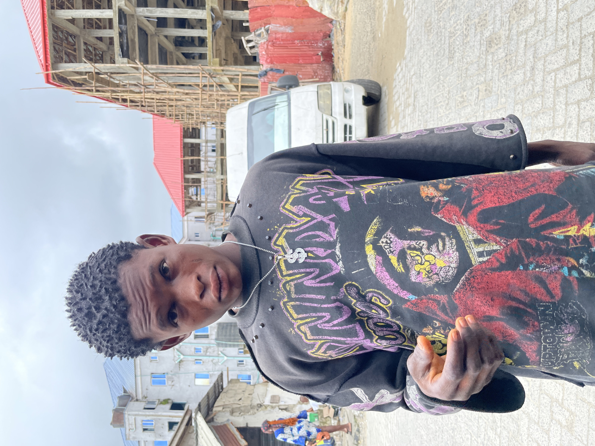 A young Black male stands outdoors, facing the camera in a medium shot. He has short, textured hair and is wearing a dark grey long-sleeved shirt with a large graphic print featuring vibrant colors and text. A silver chain with a dollar sign pendant hangs around his neck. His right hand is held up near his chest, fingers slightly splayed. The background suggests an urban setting in Ado, Nigeria, with buildings under construction, including a structure with a red roof and scaffolding. A white van is parked behind him, and other buildings and a person in a blue and white patterned dress are visible in the distance. The ground is paved with light-colored bricks. The sky is overcast.