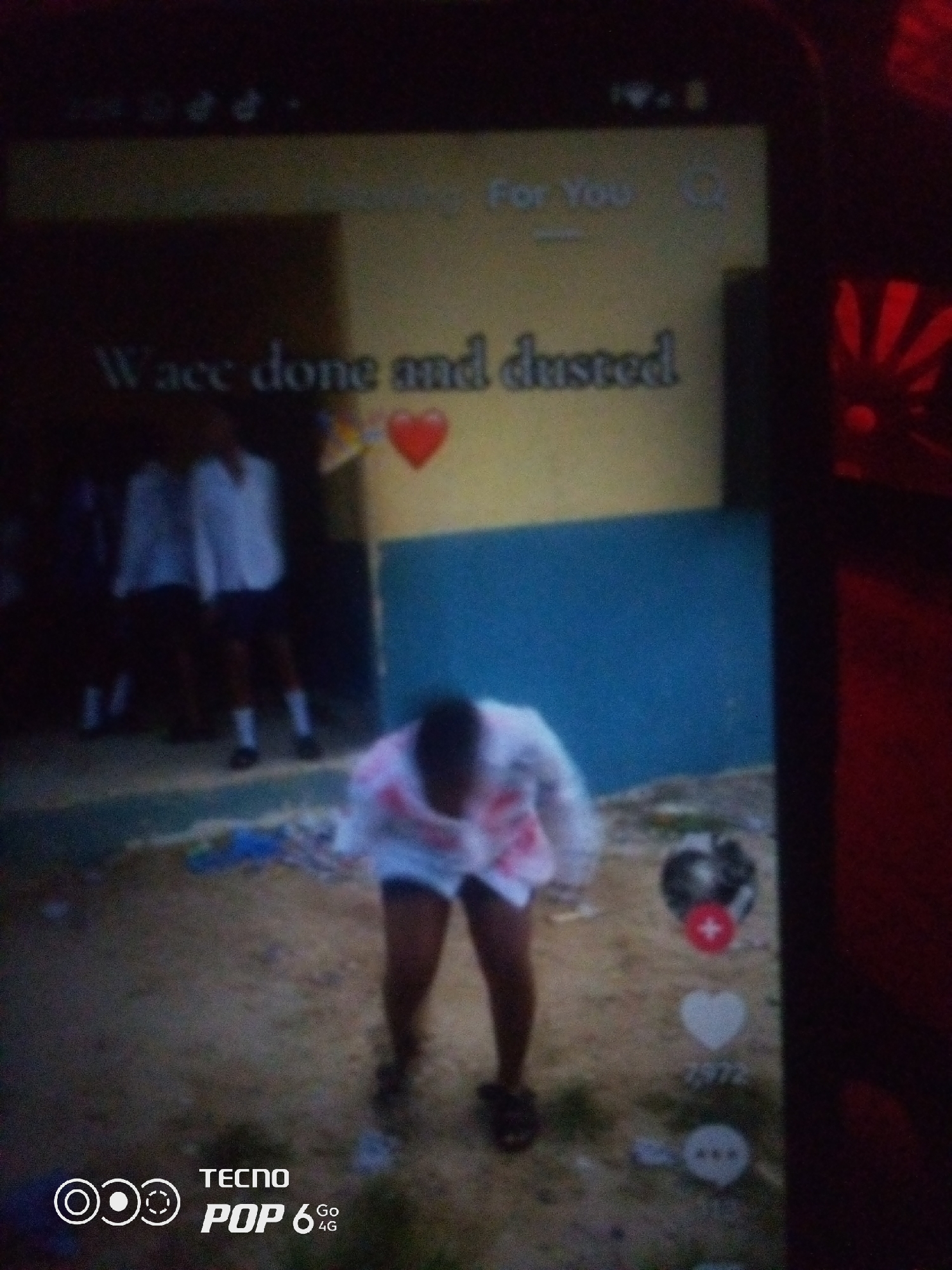 The image shows a blurry smartphone screen displaying a TikTok-like interface. On the screen, a video is playing with text overlays. The text "Wace done and dusted" is visible, along with a red heart emoji. There are also icons for likes, comments, and shares. In the video itself, a person is visible in the foreground, appearing to be dancing. In the background, several other people, possibly students in uniform, are standing. The setting appears to be an outdoor area with a building in the background, painted yellow and blue. The bottom of the screen shows the "TECNO POP 6 4G" logo. The image quality is poor due to blurriness and low light conditions. There is no specific indication or discernible detail that would allow for the identification of the location within Howayi, Nigeria.