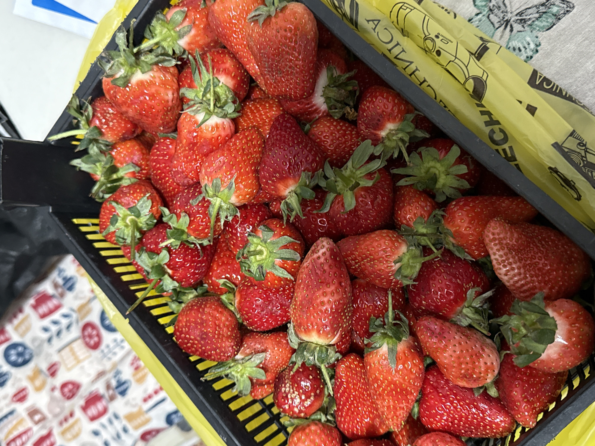 A black plastic crate filled with fresh, red strawberries with green stems sits on a surface covered with a patterned cloth. A yellow plastic bag with the word "MECHANICA" printed in reverse is partially visible along the side of the crate. The table cloth beneath the basket has a pattern of kitchen tools.
There is not enough information to determine if it is in Saint Petersburg, Russia.