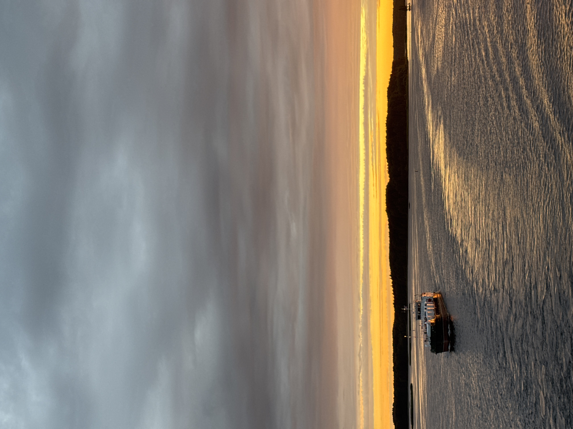A barge with white cargo containers is traversing a body of water, possibly a river or strait, at sunset. The sky is filled with dramatic clouds, transitioning from dark gray at the top to vibrant orange and yellow hues near the horizon. A dark, forested shoreline is visible in the distance, silhouetted against the colorful sky. The water reflects the warm colors of the sunset, creating streaks of light on its surface. The scene suggests a serene and atmospheric moment, likely captured in the early evening. The city of Yannovolok, Russia, is known for its waterways and natural beauty, making it a plausible location for such a scene.