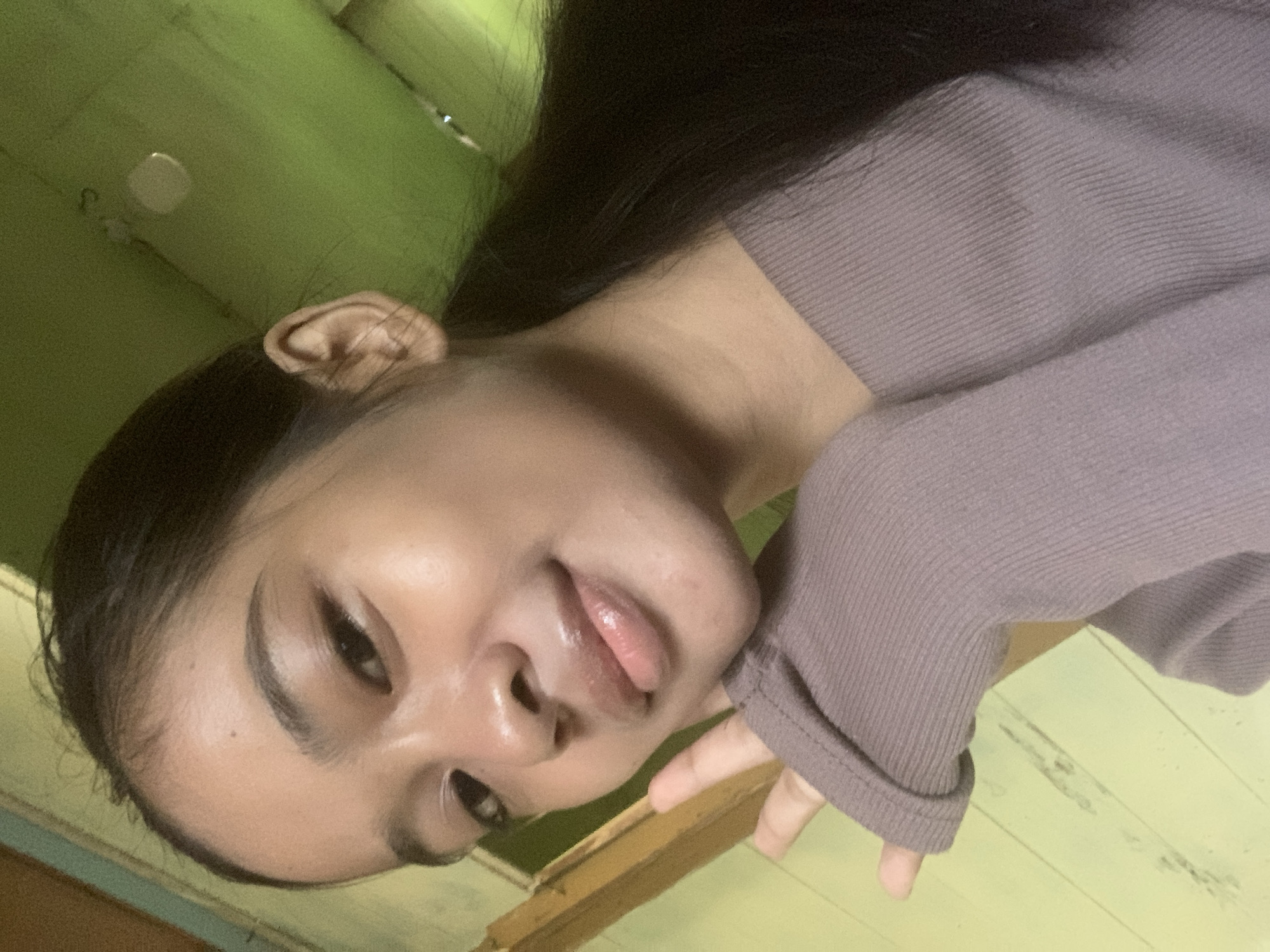 The image shows a young woman with dark hair tied in a ponytail, posing for a selfie indoors. She is wearing a mauve knitted top. Her left hand is raised, with her fingers near her face. The background consists of a light green wall with wooden paneling. There is a wooden frame on the left side of the image. The lighting suggests an indoor setting, possibly a residential room. There is no indication of the specific location within Sungai Meriam, Indonesia.