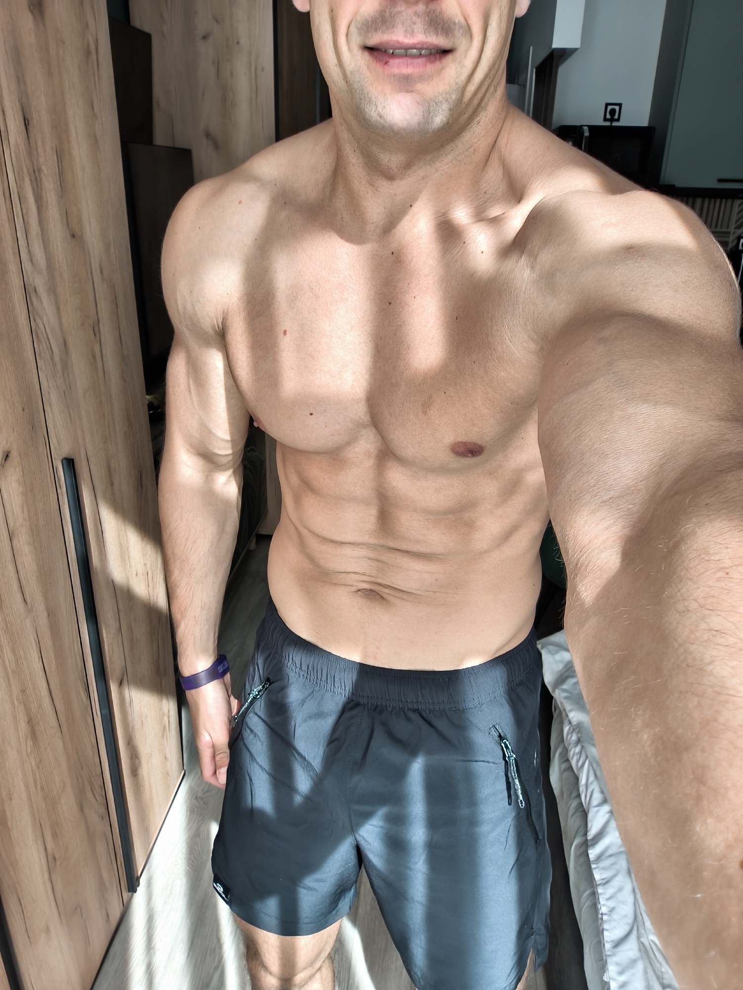 A muscular, light-skinned man is taking a selfie indoors. He is wearing dark shorts and no shirt, revealing his abdomen, chest, and arms. A purple fitness tracker is on his left wrist. He is standing in front of a wooden wardrobe. Sunlight is streaming in, creating shadows. A bed is visible on the right. The location cannot be determined to be Moscow, Russia from the image.