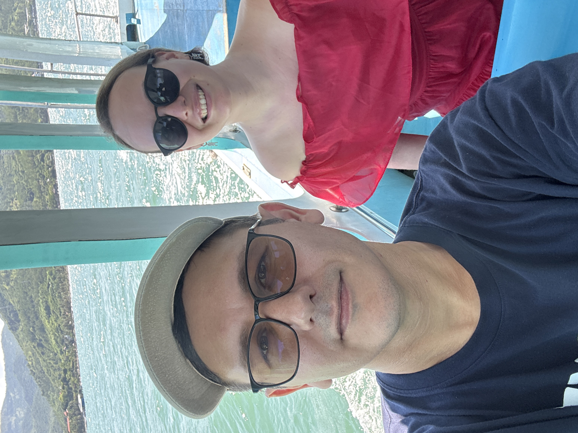 A couple is taking a selfie on a boat in Nha Trang Ward, Vietnam. The man in the foreground is wearing a dark blue t-shirt and a beige baseball cap, with glasses. The woman behind him is wearing a red off-the-shoulder top and sunglasses. They are both smiling. The sea is visible in the background, with green hills on the horizon. The boat appears to be a passenger ferry.