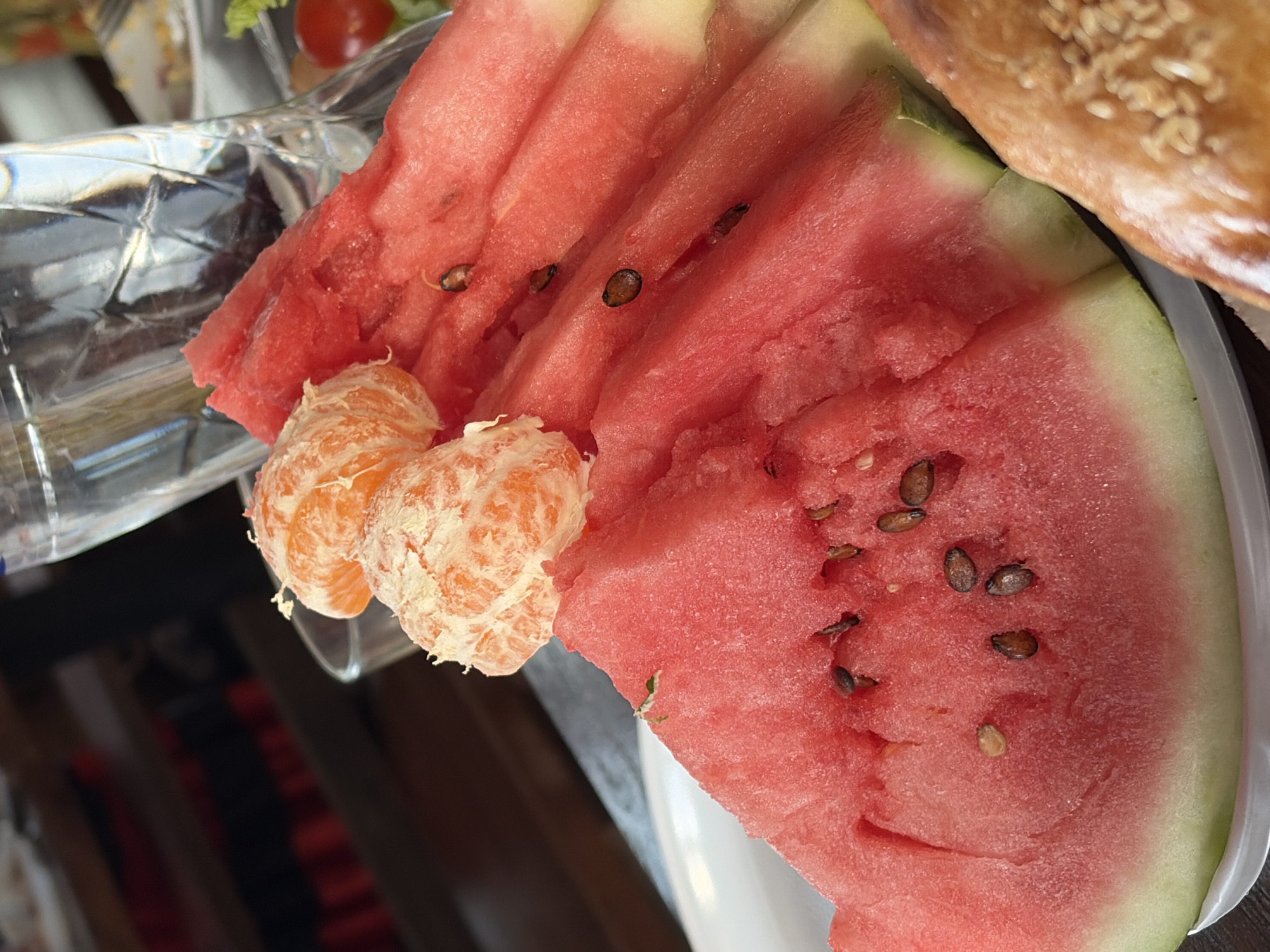 The image shows a close-up view of a plate of sliced watermelon and peeled tangerines, with a glass of water and a partially visible pastry in the background. The watermelon slices are a vibrant red with green rinds and visible black seeds. Two segments of peeled tangerine are placed on top of the watermelon. The glass of water is tall and clear, reflecting the light. A small cherry tomato and some green leaves are also visible in the background. The scene appears to be indoors, possibly at a restaurant or home, with blurred shelves in the background. The lighting suggests it might be daytime. There is no indication of the location being in the city of Chaikovsky, Russia.