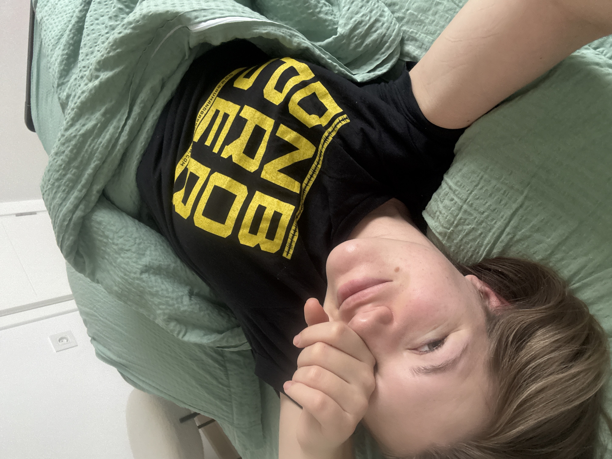 A person is lying in bed under a light green blanket. They are wearing a black t-shirt with yellow block lettering. The person's face is visible, and they are covering part of their face with their hand. The environment appears to be a bedroom, with a white wall and electrical outlet visible in the background. The location cannot be determined based on the image.