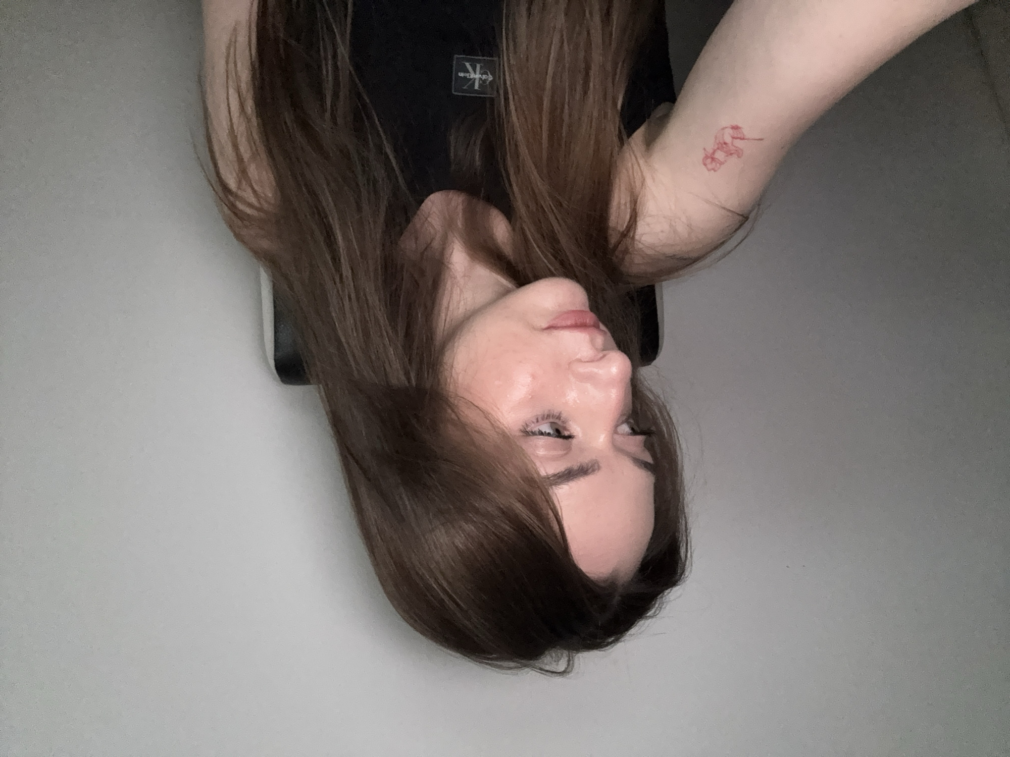 A woman with long, light-brown hair is facing the camera, though rotated 180 degrees. She is wearing a black shirt with a logo on the upper left side. Her skin is fair, and she has a small red tattoo on her upper left arm. The background is a plain, light-grey wall. There is no indication of this image being taken in Kragujevac, Serbia.
