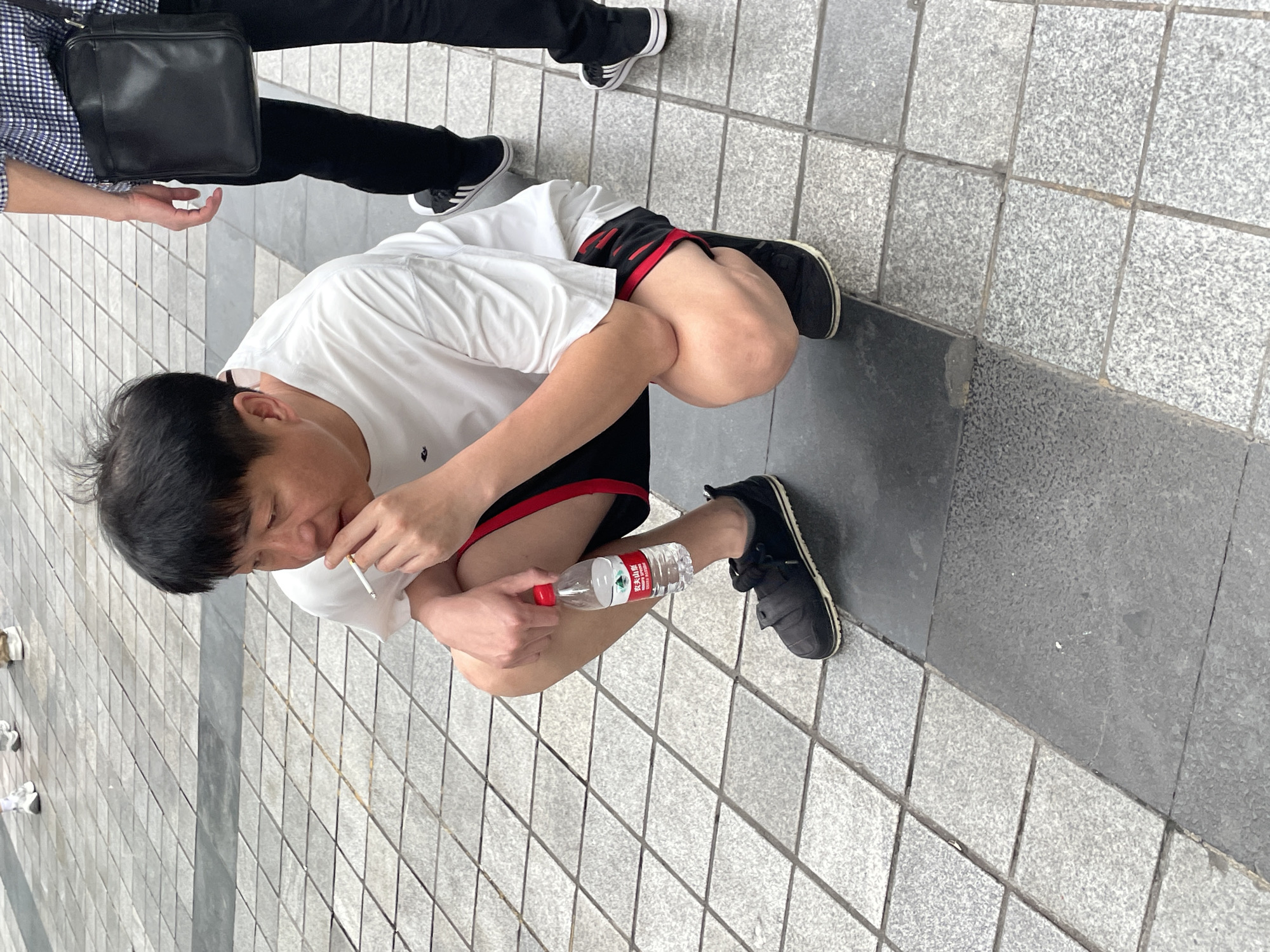 A man is squatting on a gray tiled surface, holding a cigarette in his left hand and a partially labeled water bottle in his right. He is wearing a white t-shirt, black and red athletic shorts, and black sneakers. Another person wearing black pants and black and white shoes is partially visible standing behind him. The location is likely an outdoor area with a tiled surface, which may be in the Yuzhong District, China.