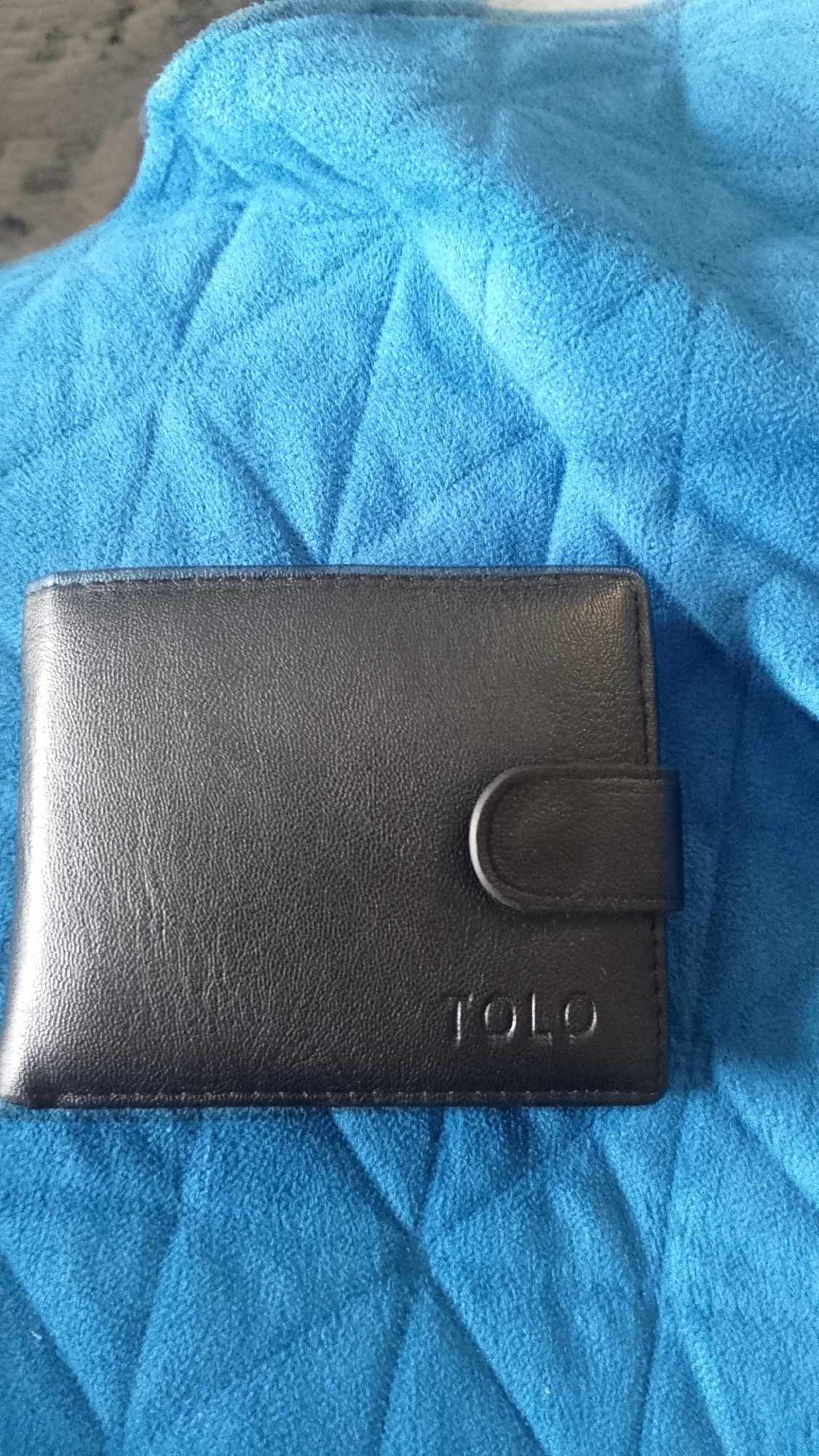 The image features a black, rectangular wallet prominently displayed on a vibrant blue, textured fabric. The wallet is made of a material that appears to be leather or a synthetic equivalent, exhibiting a finely pebbled surface. It includes a small, curved flap with a snap closure positioned on its right side. On the lower right corner of the wallet's front, the word "TOLO" is clearly embossed in capital letters.
The background fabric is a soft, fluffy textile, likely a towel or blanket, characterized by its visible loops and numerous folds and creases. Its color is a bright, uniform blue, possibly a shade of turquoise or cyan. The lighting is soft and even, highlighting the textures of both the wallet and the fabric without harsh shadows.
No people are visible in the image, and no specific activity is taking place; the wallet is stationary. The setting appears to be an indoor environment. There are no discernible cues for the time of day, weather, or emotions. The focus is entirely on the wallet and the textile it rests upon.