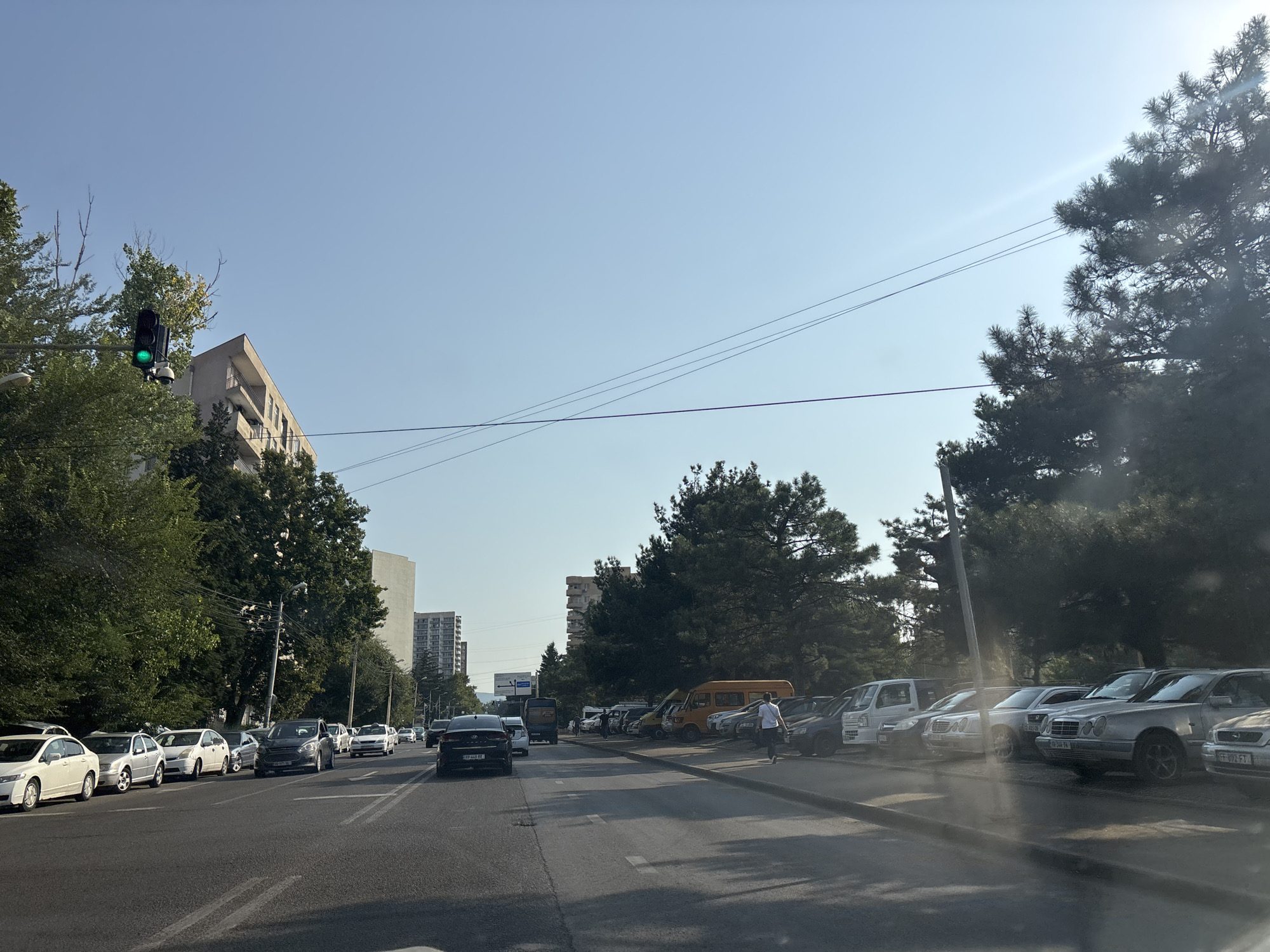 The image shows a street scene in Tbilisi, Georgia. On the left, there is a multi-lane road with cars driving and parked. A traffic light is visible with a green signal illuminated. Several cars are parked along the side of the road, including white sedans and a dark sedan. In the background, there are tall residential buildings and lush green trees lining the street. On the right side of the road, a row of parked cars, including older model Mercedes-Benz sedans and vans, are visible. A person in a white shirt and dark shorts is walking away from the parked cars. The sky is clear and blue, with a bright sun casting light and lens flares across the image, suggesting it is daytime. The overall scene depicts a typical urban environment in Tbilisi.