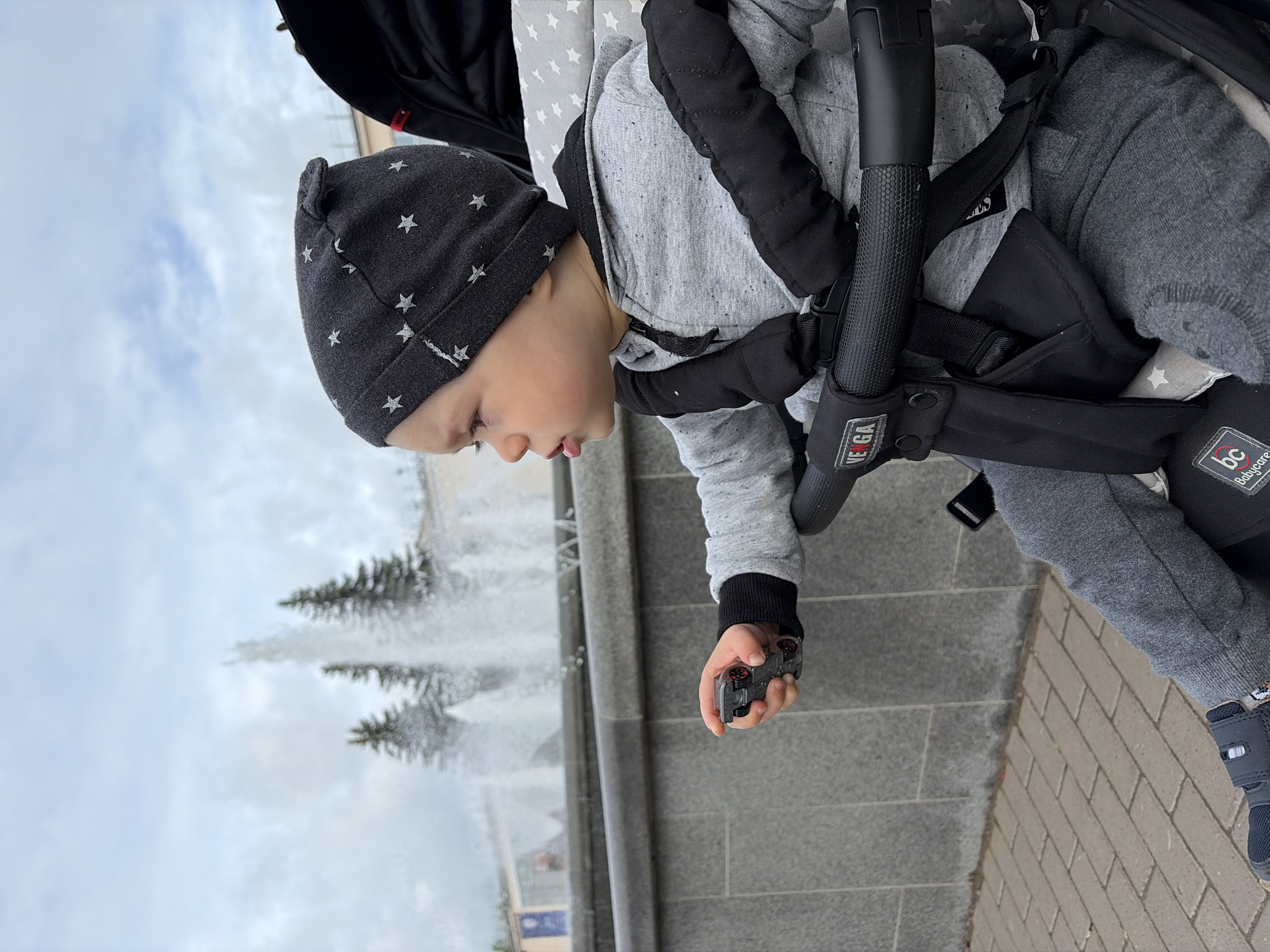 A young child is seated in a dark stroller, looking intently towards a fountain. The child wears a dark grey beanie with white stars, a light grey hooded sweatshirt, and grey sweatpants, with dark shoes featuring velcro straps. In their right hand, they hold a small, dark-colored toy car.
The scene is outdoors, likely in a park or public square in Chaikovsky, Russia. A multi-jet fountain is visible to the child's left, with water spraying upwards. Coniferous trees are blurred in the background beyond the fountain. The ground beneath the stroller is paved with light grey square tiles, transitioning to light brown brick pavers. The sky is overcast and cloudy, suggesting a daytime scene with mild or cool weather.
The child appears calm and focused, observing their surroundings while holding their toy. Visible text includes "VEGA" on the stroller's central handle and "BC Babycare" on one of the padded harness straps.
