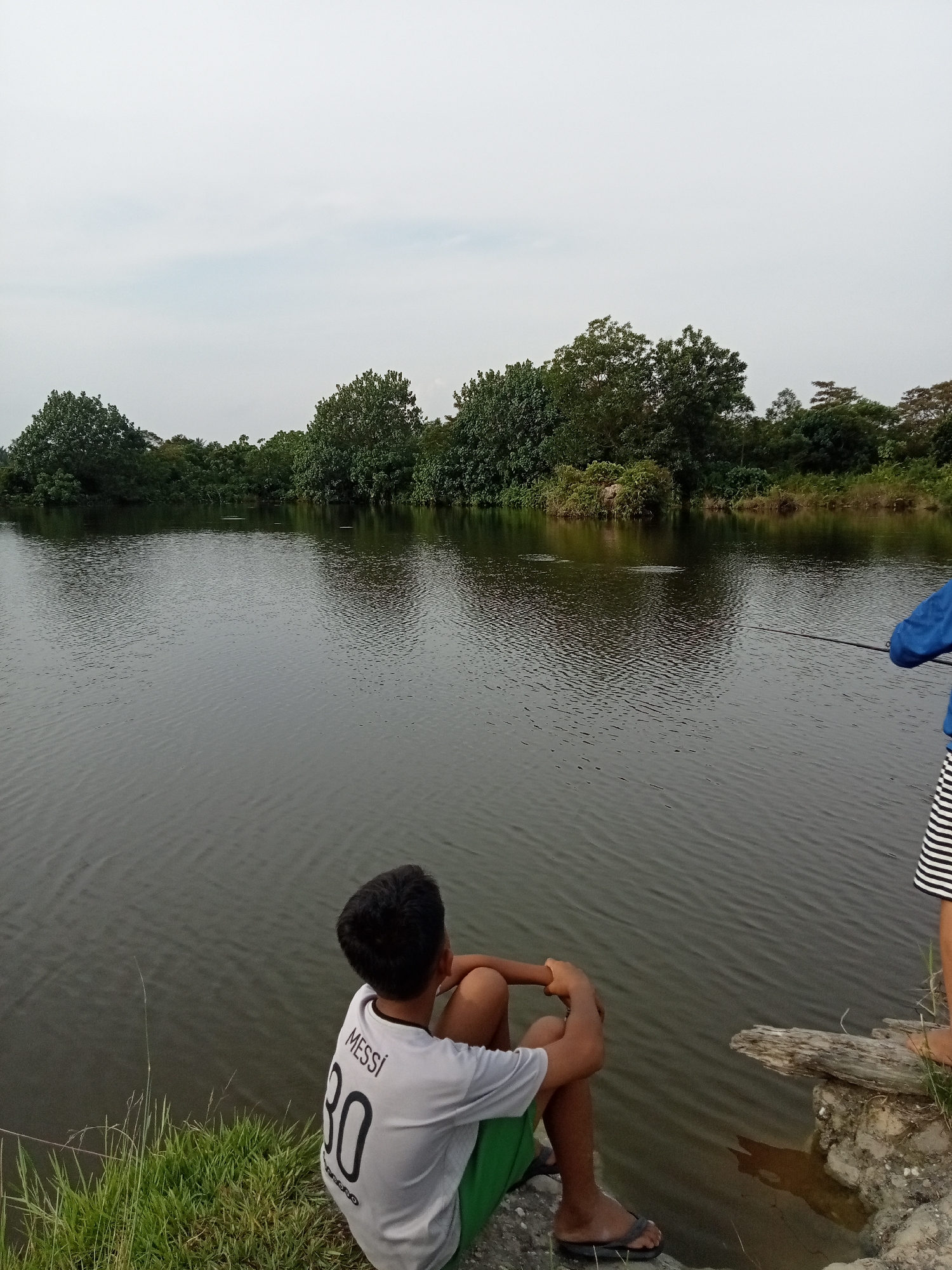 In Kualu, Indonesia, two young individuals are depicted near a body of water. In the foreground, a boy wearing a white t-shirt with "MESSI 30" printed on the back and green shorts sits on the grassy bank, facing away from the camera and towards the water. His posture suggests contemplation or observation. To the right, another individual, partially visible, is standing and holding a fishing rod, engaged in fishing. The water is calm with ripples, and the background features a lush, green treeline lining the opposite bank of the water. The sky is overcast. The scene appears to be a tranquil outdoor setting, possibly a lake or river, in a natural or semi-natural environment within Kualu.