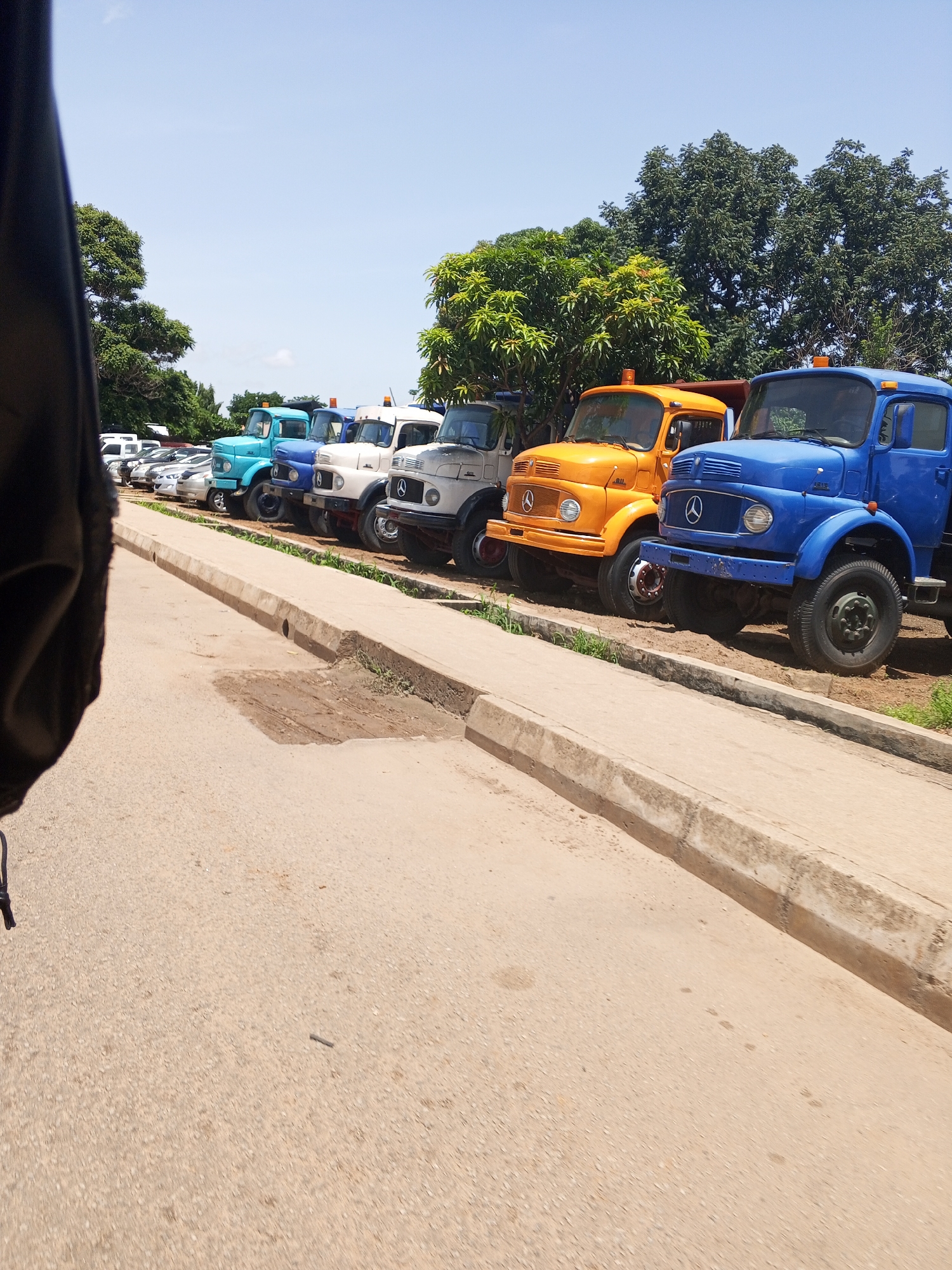 A line of parked trucks, primarily dump trucks and a few smaller vehicles, are visible from a low-angle perspective. The trucks are predominantly older models, with a variety of colors including blue, orange, and white. Some of the trucks have what appear to be orange hazard lights on their roofs. The vehicles are parked on a dirt or gravel lot adjacent to a paved road with a concrete curb. In the background, lush green trees provide a backdrop, and the sky is clear and blue. The foreground shows the textured surface of the paved road and the curb. The image appears to have been taken from within another vehicle, as evidenced by the dark, blurred object on the left edge of the frame. There are no people visible in the image. The scene suggests a storage or parking area for vehicles in a locale with abundant vegetation.