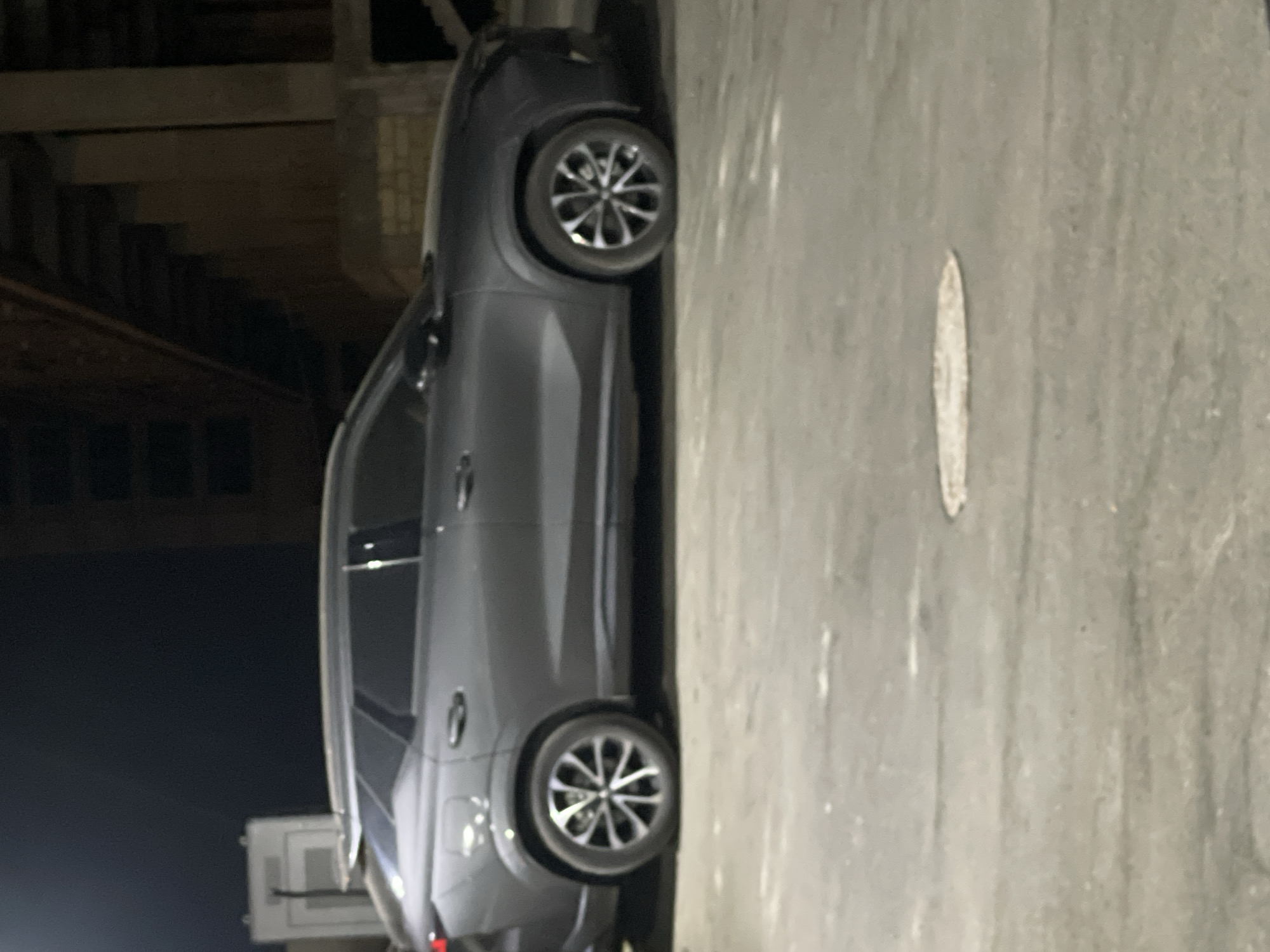 A gray SUV is parked on a paved surface at night, possibly an urban area. The vehicle is seen from its side profile, showing both the front and rear wheels. The wheels have a distinctive multi-spoke design. In the background, there are structures that suggest an industrial or construction site, with visible concrete pillars and beams. The lighting indicates it is nighttime. The image was taken in Sumgayit, Azerbaijan.