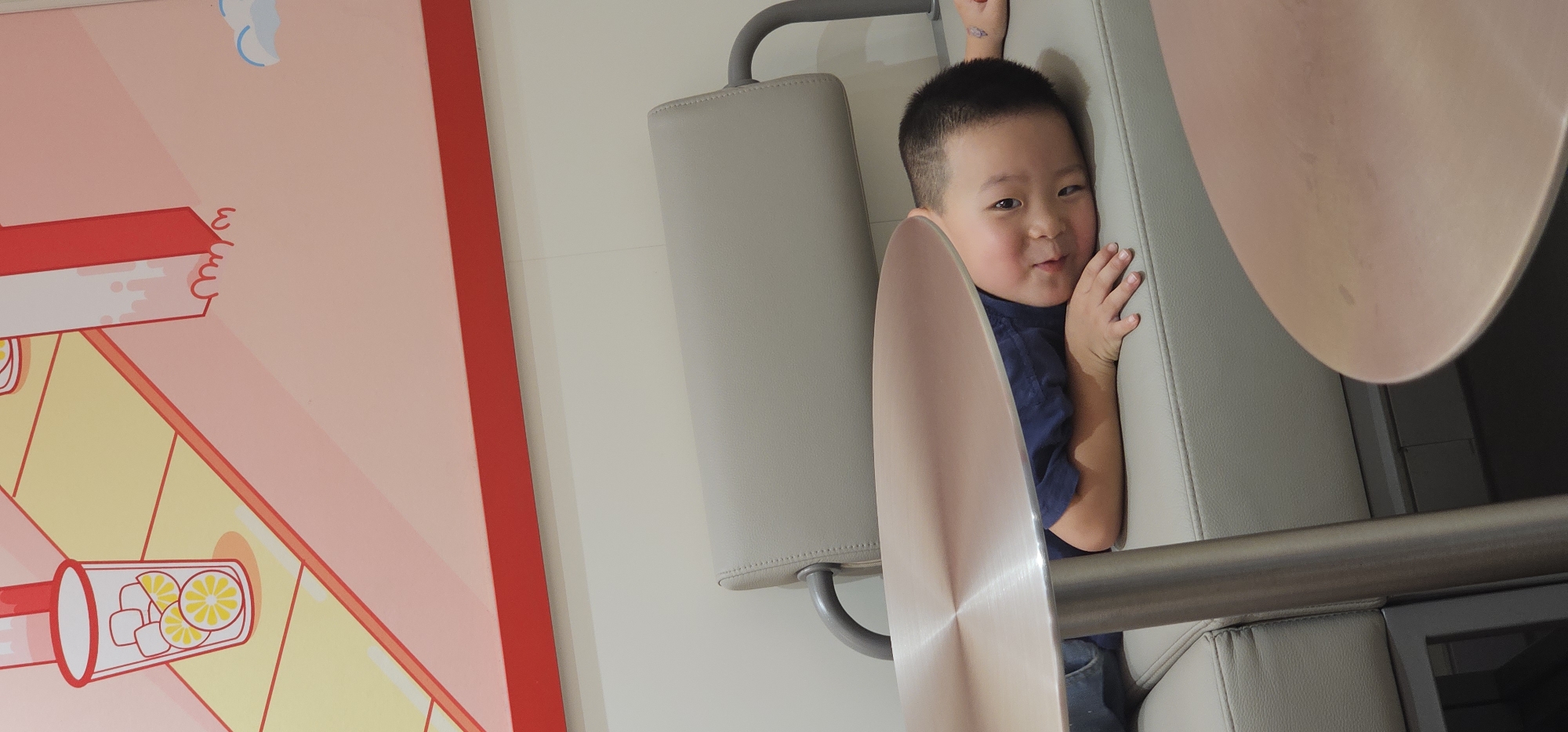 A young East Asian boy with short dark hair is lying on his side on a light grey couch, with his head propped on his arm. He is wearing a dark blue t-shirt and blue pants. His eyes are open, and he is looking towards the camera with a slight smile. A part of a round, rose-gold colored table is in the foreground, obscuring the boy's torso. The background consists of a light grey wall and a large, colorful mural featuring stylized drinks and geometric shapes in pink, yellow, and red. The image appears to be taken indoors, possibly in a cafe or waiting area. The boy's position suggests he is relaxed or possibly resting. The presence of the mural and the furniture indicates a public or semi-public space. There are no other people visible in the frame. The lighting is moderate, casting soft shadows.