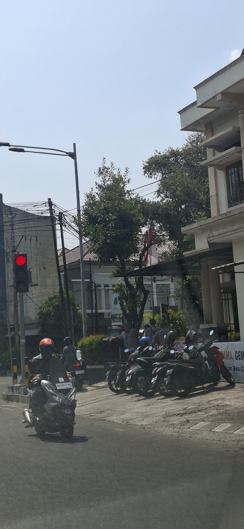A motorcycle is parked on the left side of the road, with its rider wearing a red helmet and a blue mask. Several other motorcycles are parked in a lot to the right, adjacent to a building. Some people are gathered near the building's entrance. The red light of a traffic signal is illuminated. The scene appears to be in a moderately urban area, with overhead power lines visible and trees lining the street. A banner with Indonesian text is partially visible on the building.