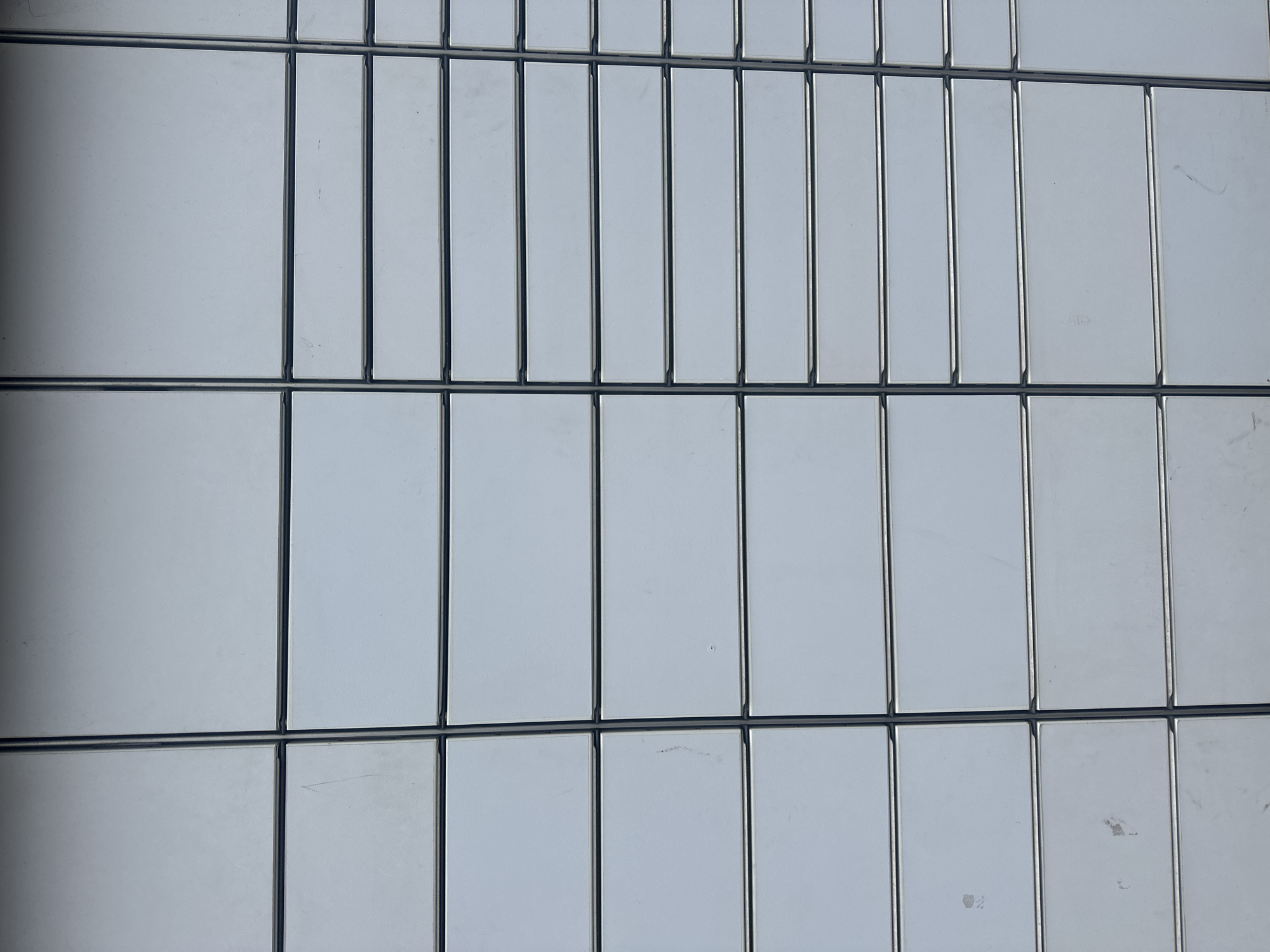 The image shows a close-up of a building facade with a grid-like pattern. The facade is composed of rectangular, light-colored panels divided by dark, narrow strips. The panels appear uniform in color and texture. The grid pattern is horizontally and vertically consistent, forming rows and columns of panels. There is no indication of the location being Tunbridge Wells, United Kingdom. No people, subjects, actions, or interactions are visible in the image.