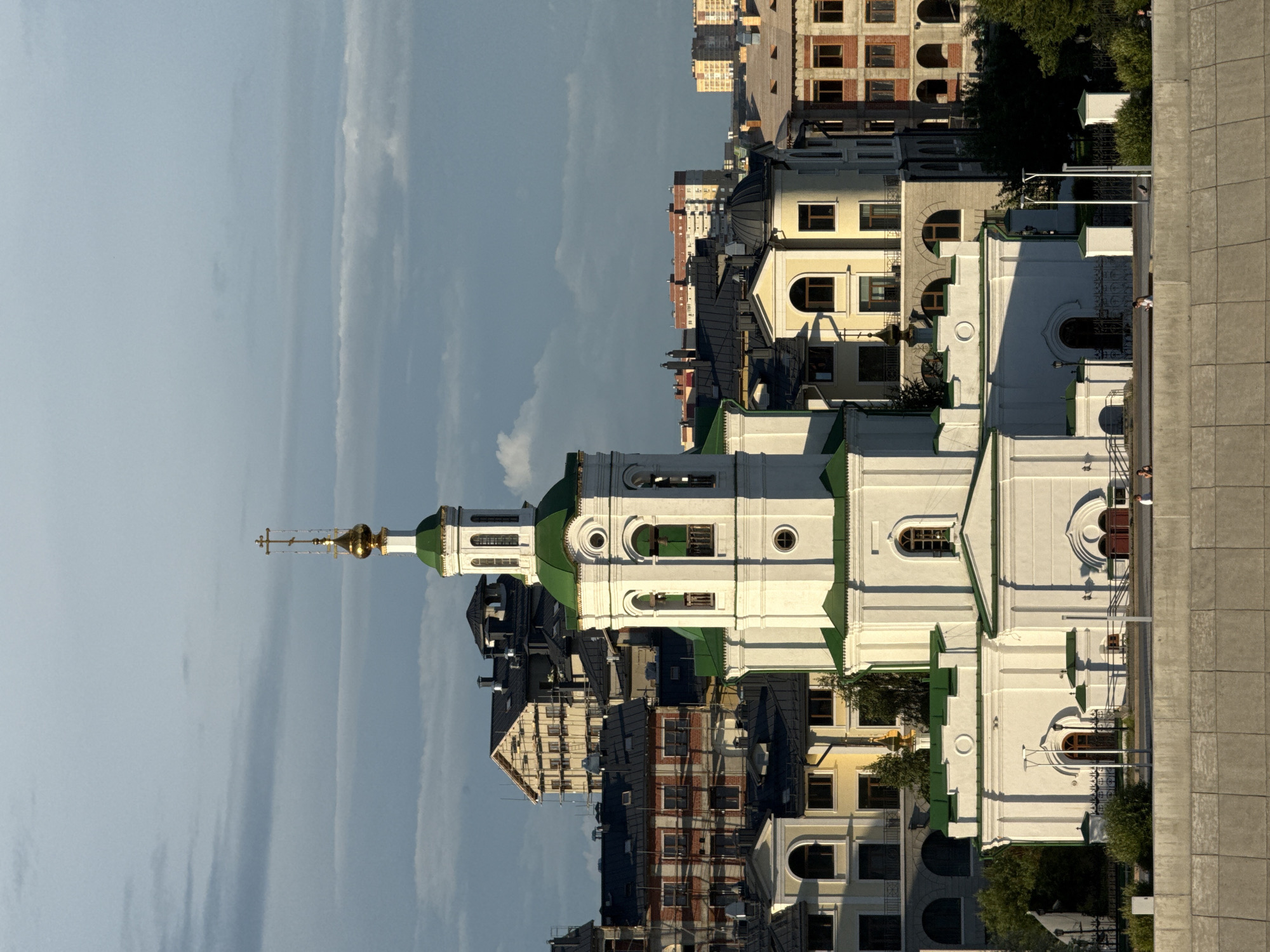 The image shows a view of the Church of the Assumption of the Holy Virgin in Tyumen, Russia. The church is a white, multi-domed structure with a tall bell tower topped by a golden dome and cross. It is situated on a waterfront, with a concrete embankment in the foreground. Behind the church, a cityscape of various buildings, including residential buildings and constructions, is visible under a cloudy sky. Several people are seen walking and sitting along the embankment. The scene appears to be captured during the late afternoon or early evening, as indicated by the lighting.