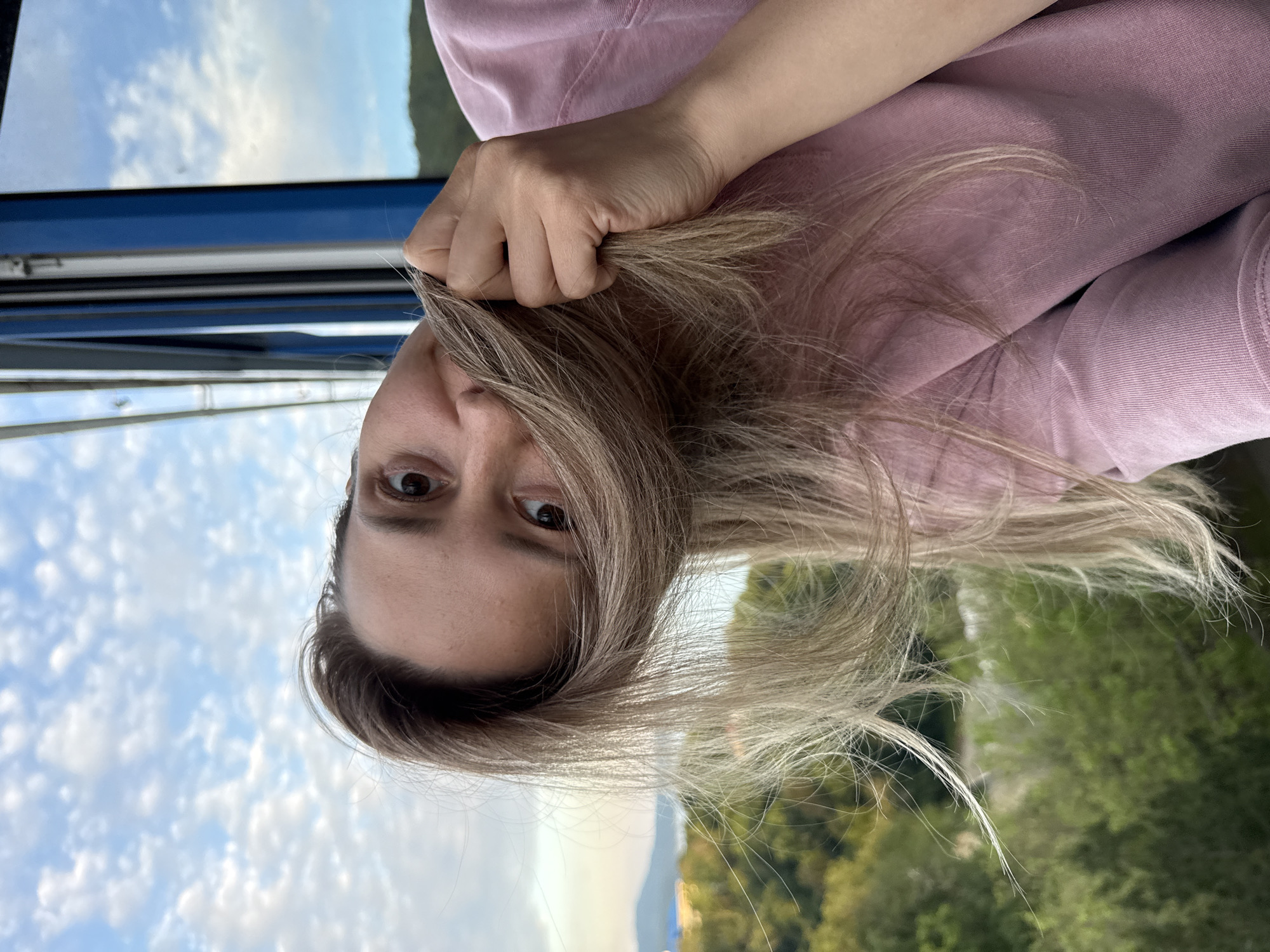 The image shows a close-up, tilted selfie of a woman with blonde hair, wearing a pink t-shirt. Her hair is partially obscuring her face, and she is holding a section of her hair with her right hand. The background suggests an outdoor setting with green trees and hills visible in the distance. The sky above is blue with scattered clouds. A blue frame, possibly from a window or cabin, is visible on the right side of the image. The scene appears to be a candid moment captured during the day. The specific location in the city of Nebug, Russia, cannot be determined from the visual information alone.