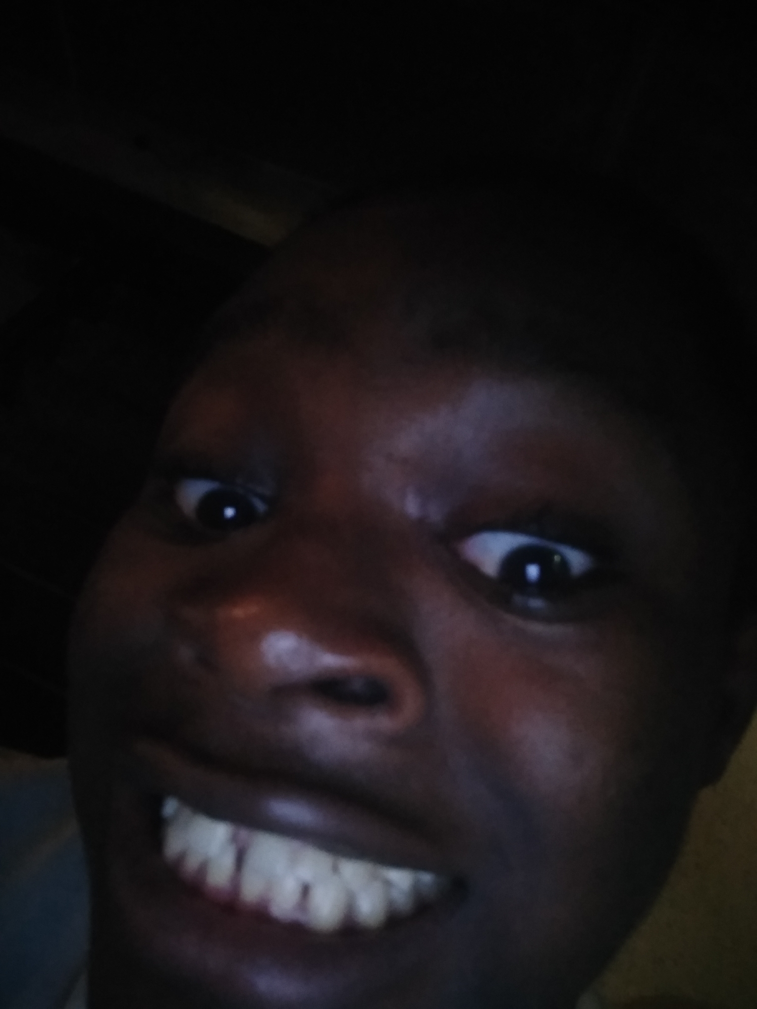 A close-up selfie of a dark-skinned person with wide eyes and a broad smile revealing teeth. The image is dimly lit. The specific location within Shangisha, Nigeria, is not identifiable. No other people or objects are visible.