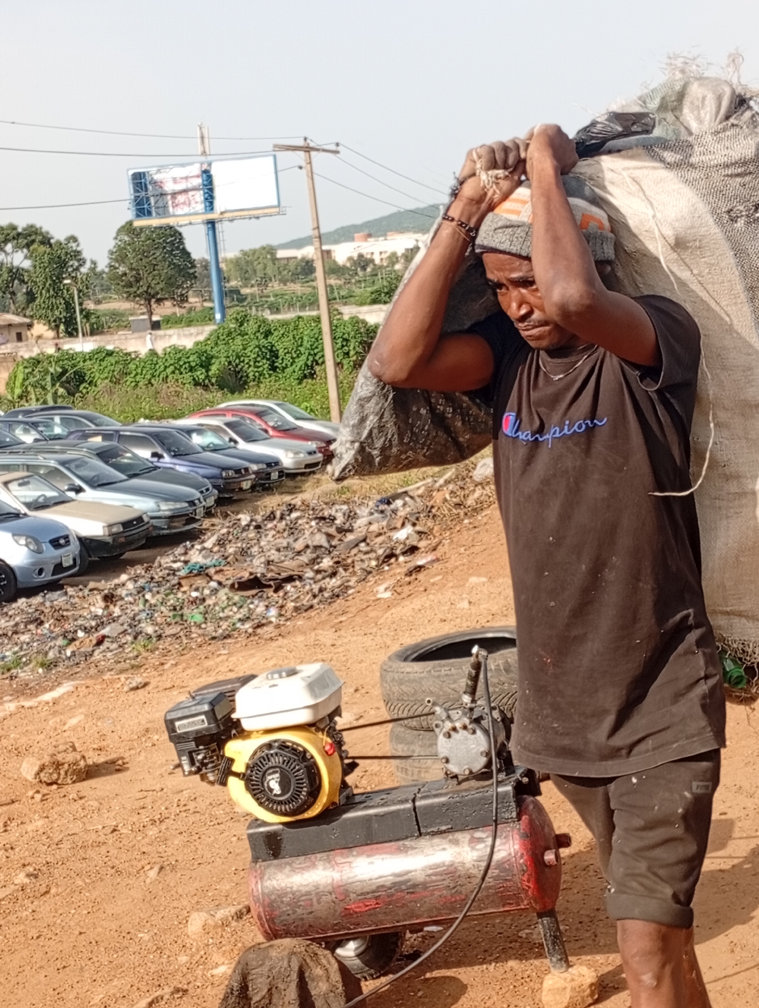 In Jos, Nigeria, a man is captured carrying a large sack over his shoulders. He is wearing a dark t-shirt with a "Champion" logo and a beanie. In the foreground, a small generator with a yellow engine is visible. Behind him, a row of parked cars lines a dusty area. The background shows a billboard and a landscape with trees and buildings under a clear sky. The scene appears to be an outdoor, possibly industrial or waste-disposal area, given the debris and the presence of the generator.