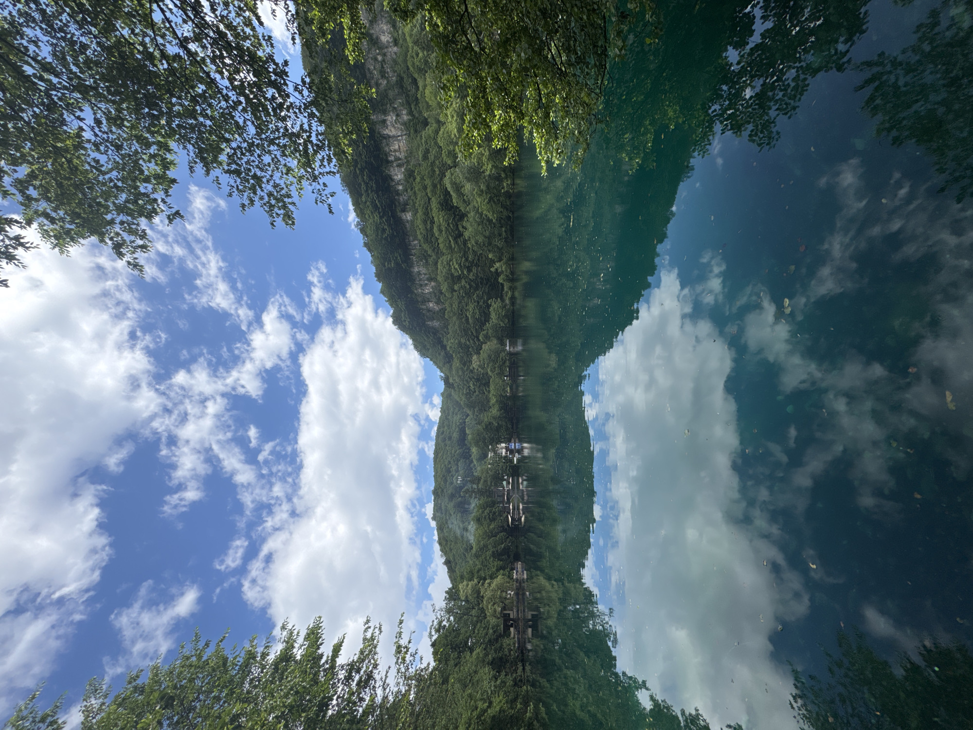 This media file captures a serene natural scene, likely taken on a clear, sunny day. The image is framed from below, looking upwards at a vibrant blue sky dotted with fluffy white clouds. Lush green trees frame the top and bottom edges of the photo, their leaves creating a dense canopy. The focal point of the image is a tranquil body of water, possibly a lake or river, that reflects the sky and the surrounding landscape with remarkable clarity. The reflection creates a mirror image, making it difficult to discern the actual orientation of the scene.
The water's surface is a deep, cool turquoise, further enhanced by the reflections of the sky and the heavily wooded mountains that rise steeply in the background. These mountains are covered in a thick carpet of green trees, with a rocky cliff face visible on the left side. The reflection of the mountains in the water is almost perfect, creating a sense of depth and symmetry. Several small structures, possibly cottages or platforms, are visible along the waterline of the mountains, hinting at human presence in this otherwise wild setting. The overall mood of the image is one of peace and natural beauty, inviting the viewer to immerse themselves in the tranquility of the location. There is no visible text in the image.
