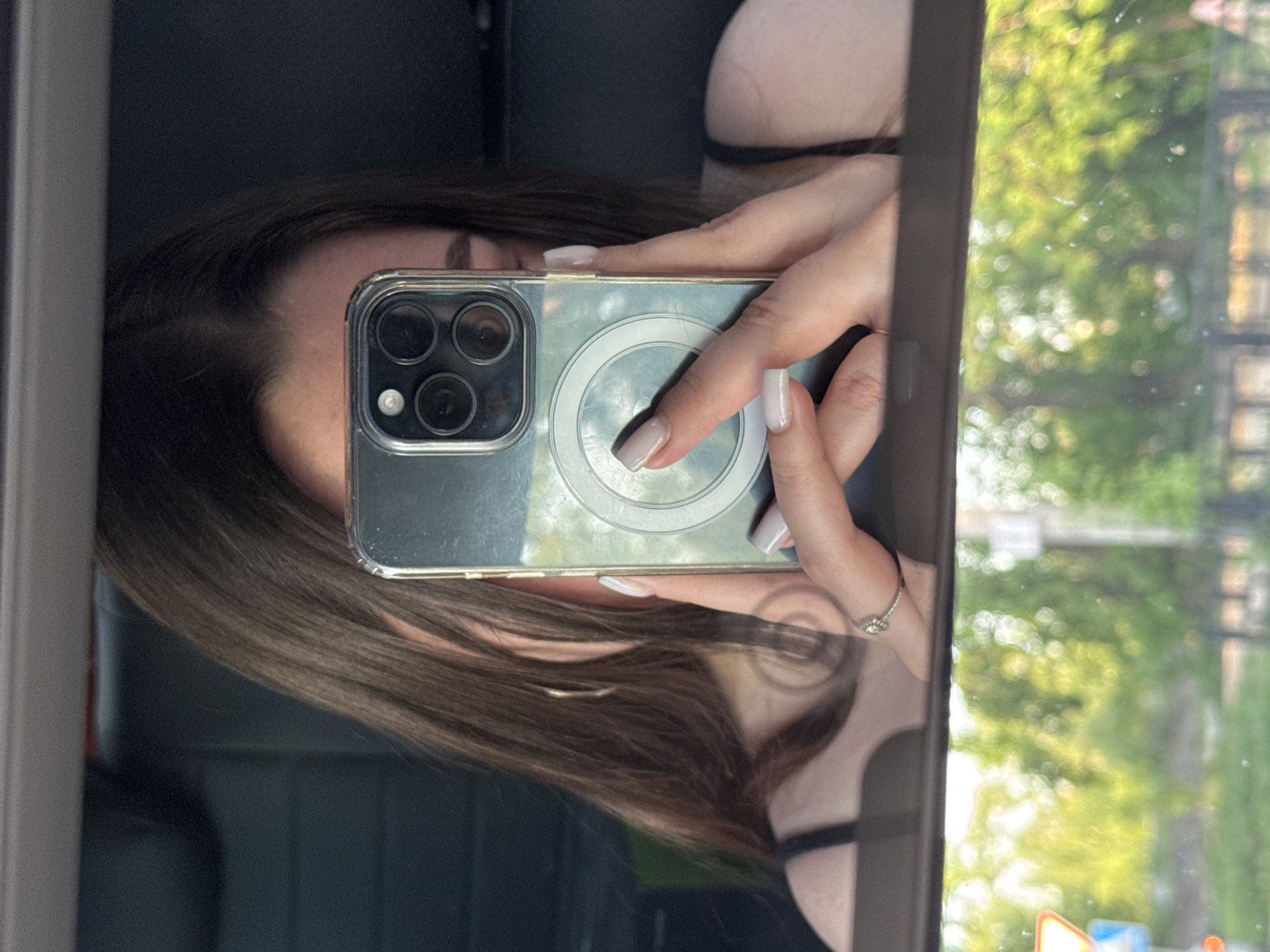 A woman is taking a selfie in what appears to be a car mirror. She is holding a phone with a clear case and a MagSafe charging accessory on the back. Her hand is visible, adorned with a ring on her ring finger, and her nails are painted. Her brown hair is partially visible. The reflection in the mirror shows trees and possibly buildings, suggesting an urban setting. A road sign is visible in the reflection.
Although the image does not confirm the location is in Perm, Russia, the presence of a road sign in the reflection might help confirm the exact location.