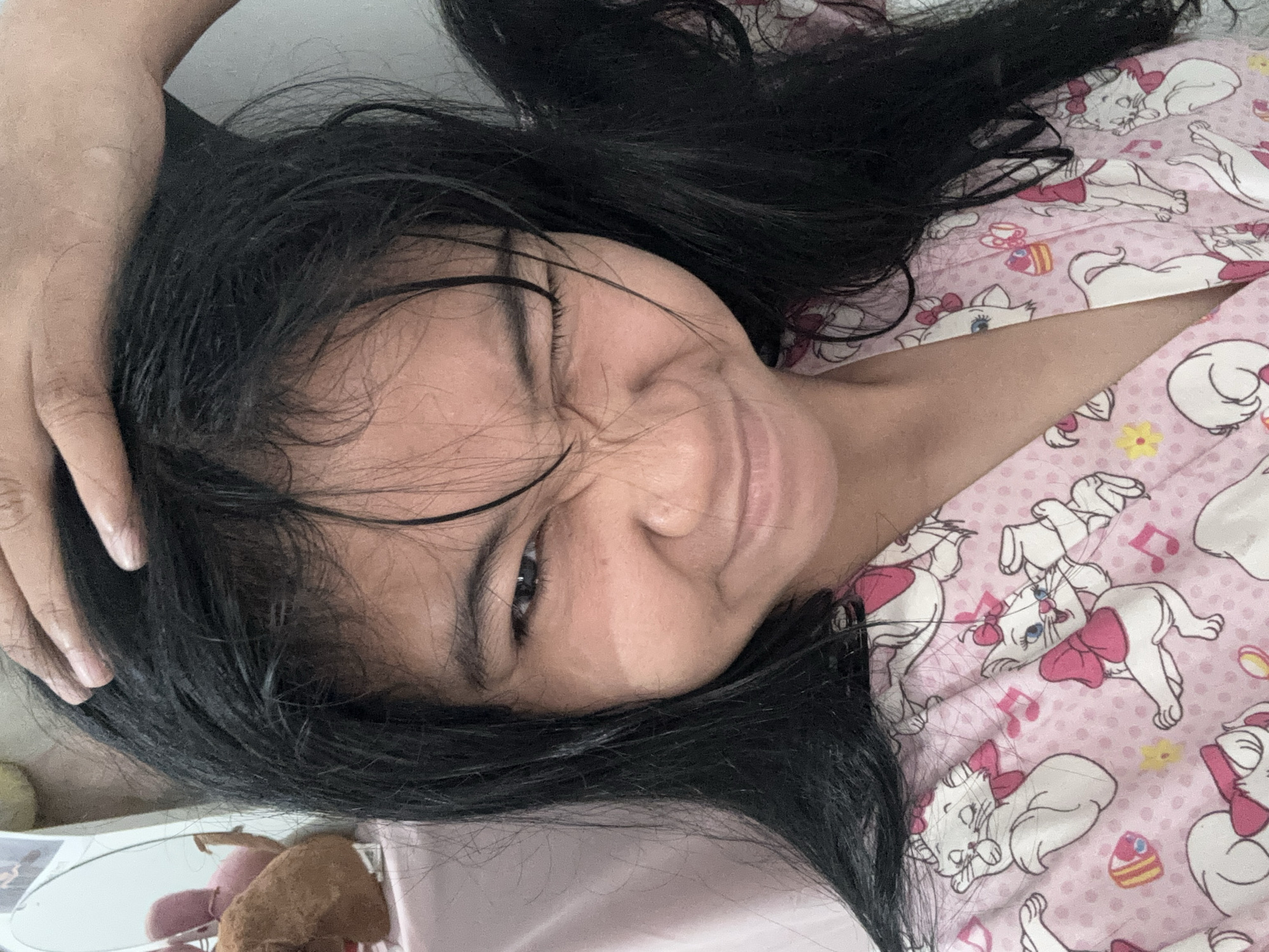The image is a close-up selfie of a young girl with long, dark hair and fair skin. She is wearing a pink pajama top with a pattern of white cats and musical notes. Her hair is partially covering her face, and she is winking with her right eye closed, while her left eye is open and looking at the camera. Her hand is resting on top of her head, seemingly pulling her hair. The background is slightly out of focus, showing a pink surface and some stuffed toys on the left. There is no visible indication of the location within North Jakarta, Indonesia.