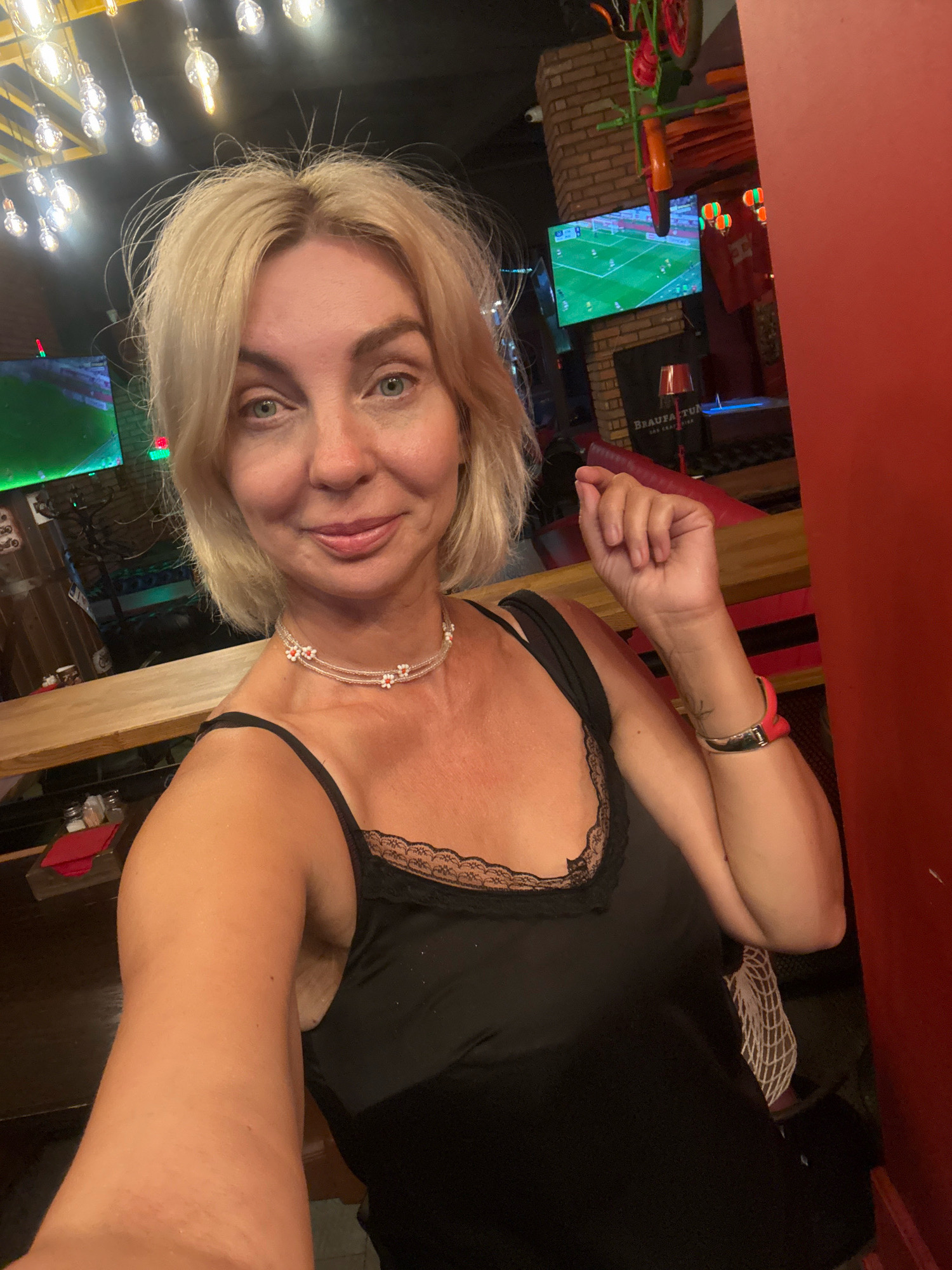 A blonde woman with blue eyes and fair skin is taking a selfie in a dimly lit, indoor establishment, possibly a bar or restaurant. She is wearing a black camisole with lace trim and a delicate beaded necklace with floral accents. Her left arm is raised, capturing the selfie. In the background, there are several televisions displaying what appears to be a soccer match, brick walls, and exposed light bulbs hanging from the ceiling. A red wall is visible to the right of the frame. The location is identified as Sochi, Russia.