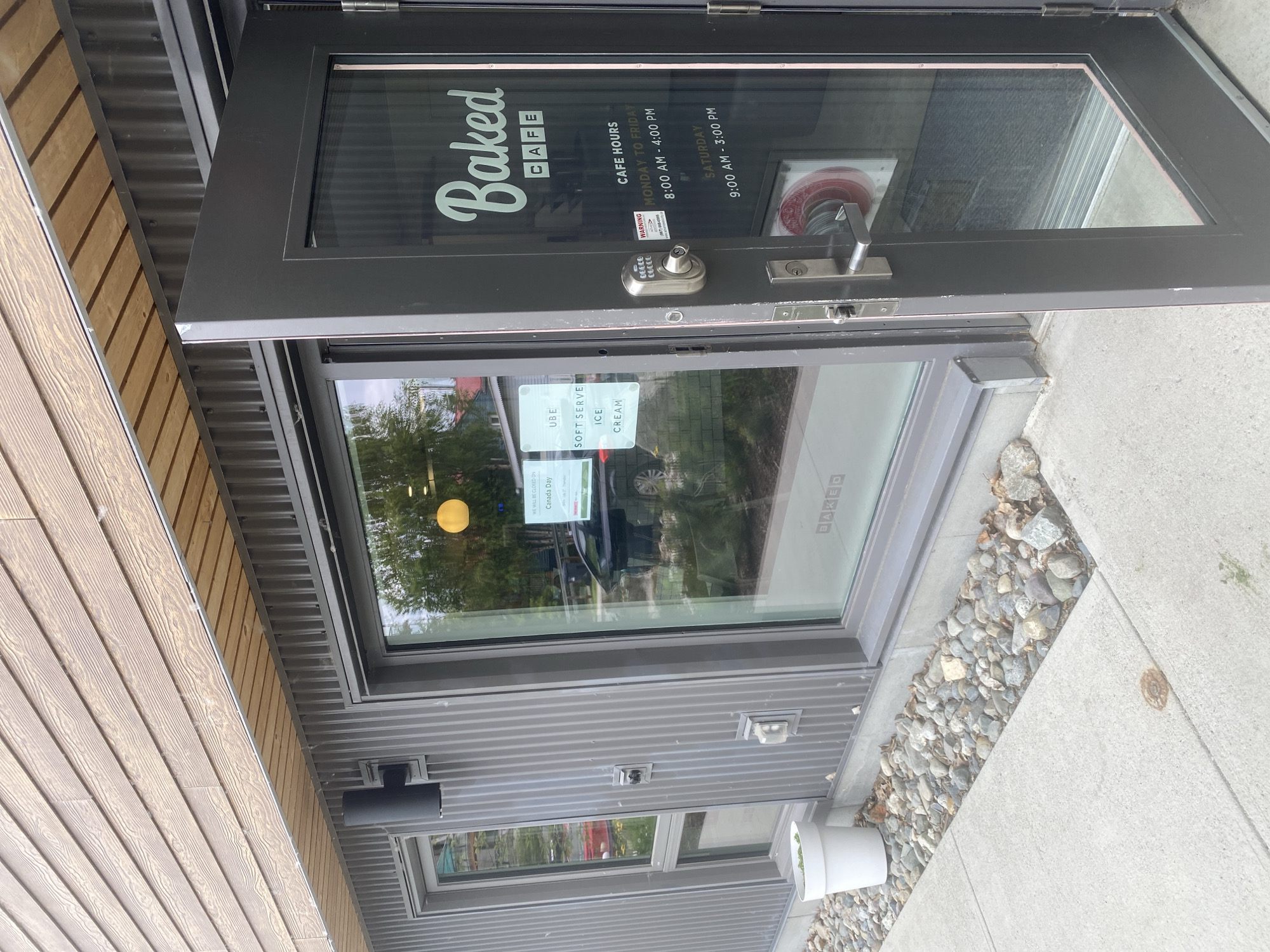 The exterior of a business called "Baked CAFE" is shown in Whitehorse, Canada. A dark gray door is ajar, revealing a glass entrance. The door has a silver digital lock and a silver handle. On the glass of the door, the business name "Baked CAFE" is written in white lettering, along with the cafe hours: Monday to Friday, 8:00 AM to 4:00 PM, and Saturday, 9:00 AM to 3:00 PM. A "WARNING" sticker is visible on the door.
To the left of the door is a large window with signage displayed inside. One sign states "WE WILL BE CLOSED ON Canada Day." Another sign reads "UBE SOFT SERVE ICE CREAM." A yellow circular light fixture is visible through the window. The building has corrugated metal siding and a wooden soffit. A black outdoor light fixture is mounted to the siding. A white planter with a plant is positioned near the window. The ground in front of the building is paved with concrete and bordered by rocks and some fallen leaves.