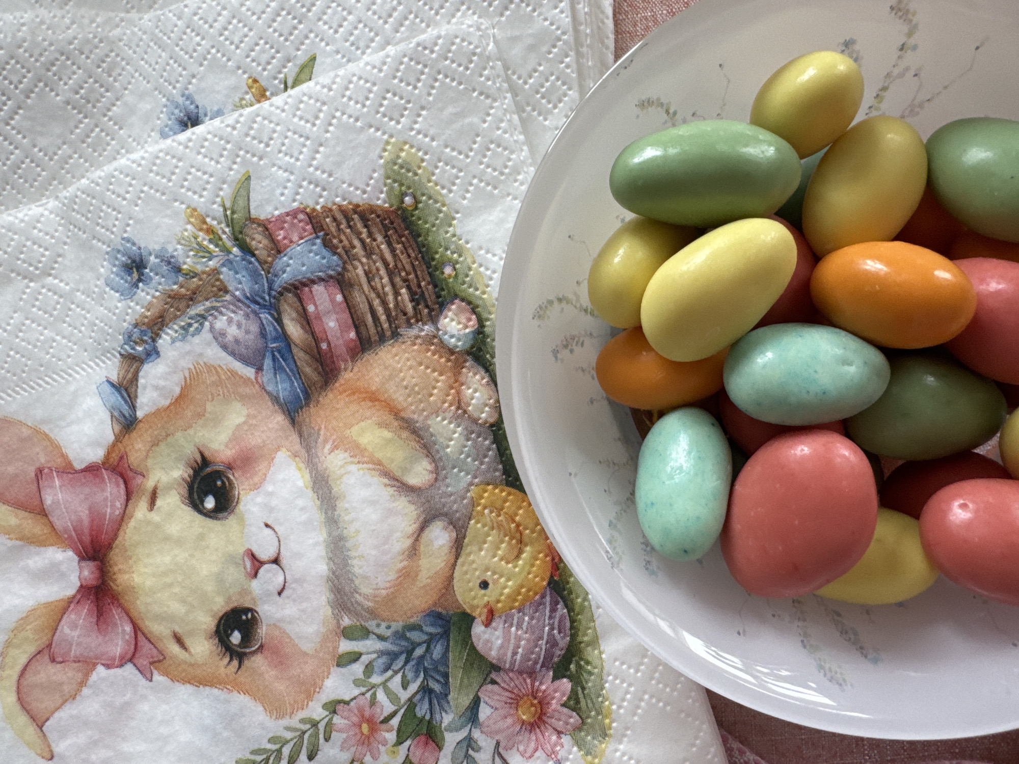 A bowl of multi-colored candy eggs sits beside a stack of printed paper napkins featuring a bunny and Easter motifs. The candy eggs are in various colors including yellow, green, orange, blue, and pink. The bunny on the napkins has a pink bow. The environment appears to be indoors. There is no information available to determine the location of Вассята, Russia.