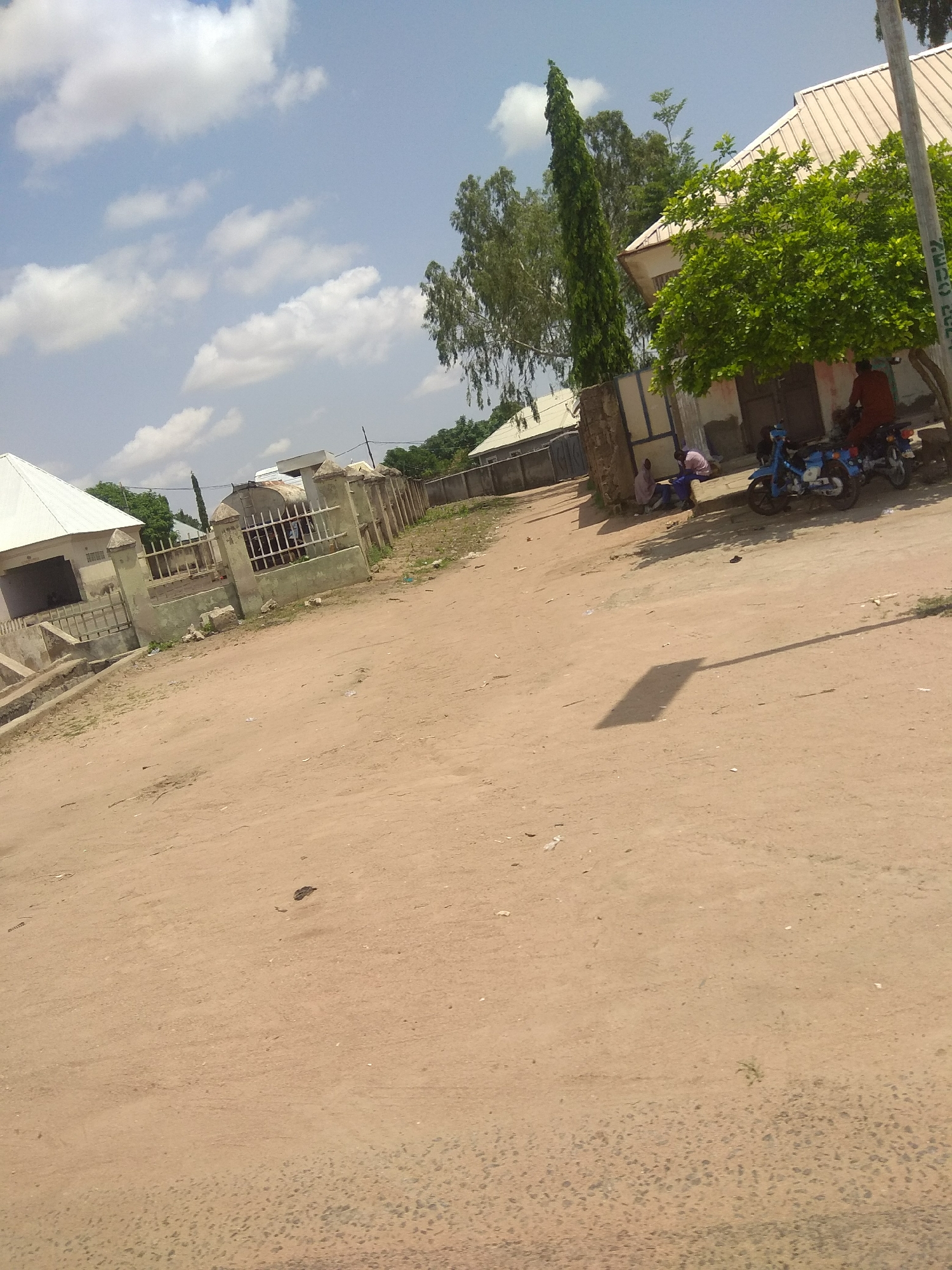 The image depicts a street scene in Jalingo, Nigeria. The foreground is dominated by a dusty, sandy road, with a clear shadow of a utility pole cast across it. In the middle ground, several structures are visible, including buildings with light-colored roofs and a fence. A tall, slender evergreen tree stands prominently behind the fence, adding to the scenery.
On the right side of the image, two motorcycles are parked. Near the motorcycles, two individuals are seated on the ground, appearing to be engaged in conversation or resting. Another person is seated on one of the motorcycles, facing away from the camera. A large, leafy green tree is situated to the right of these individuals and motorcycles, providing shade.
The background shows more buildings and vegetation, suggesting a semi-urban or rural environment. The sky is blue with scattered white clouds, indicating fair weather and likely daytime. The overall impression is one of a quiet, ordinary day in a community. There is no visible text in the image.