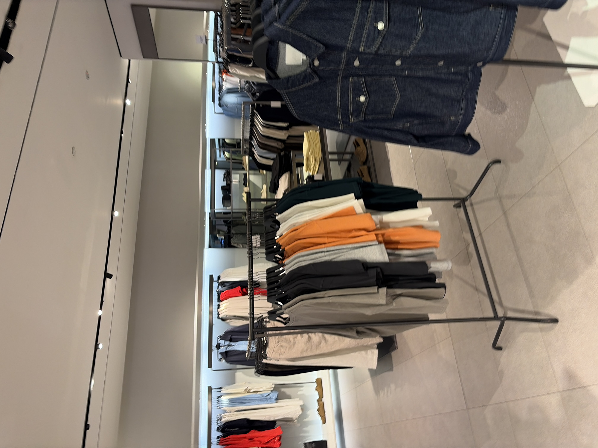 In the city of Kuta, Indonesia, this image captures the interior of a clothing store. The scene is dominated by racks of clothing, showcasing a variety of garments. On the left, a rack displays brightly colored tops in red and white, alongside pale blue and white shirts, and a black garment. Further to the left, a pair of tan shoes are visible on a shelf.
Moving towards the center, a rack presents a collection of neutral-toned clothing, including beige pants, and t-shirts in gray, black, and light gray. To the right of these, a rack holds vibrant orange and white t-shirts.
The foreground features a dark blue denim jacket hanging on a black clothing rack. Behind it, shelves are visible with stacks of yellow folded garments. The store environment is clean and well-lit, with a neutral-toned tiled floor and overhead track lighting. The background shows more clothing displays and shelving units. There are no people visible in the image.