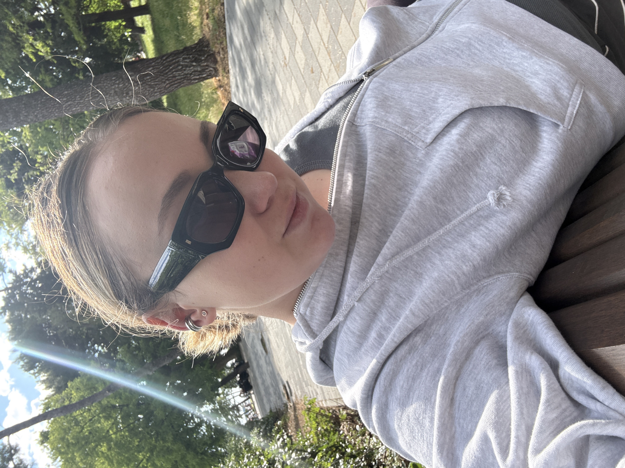 The photograph shows a woman outdoors, likely in a park or green space in Bar, Montenegro. She is wearing sunglasses and a light gray hooded sweatshirt, and is seated on a bench. The background includes trees and paving stones, suggesting a public area. The lighting indicates a sunny day.