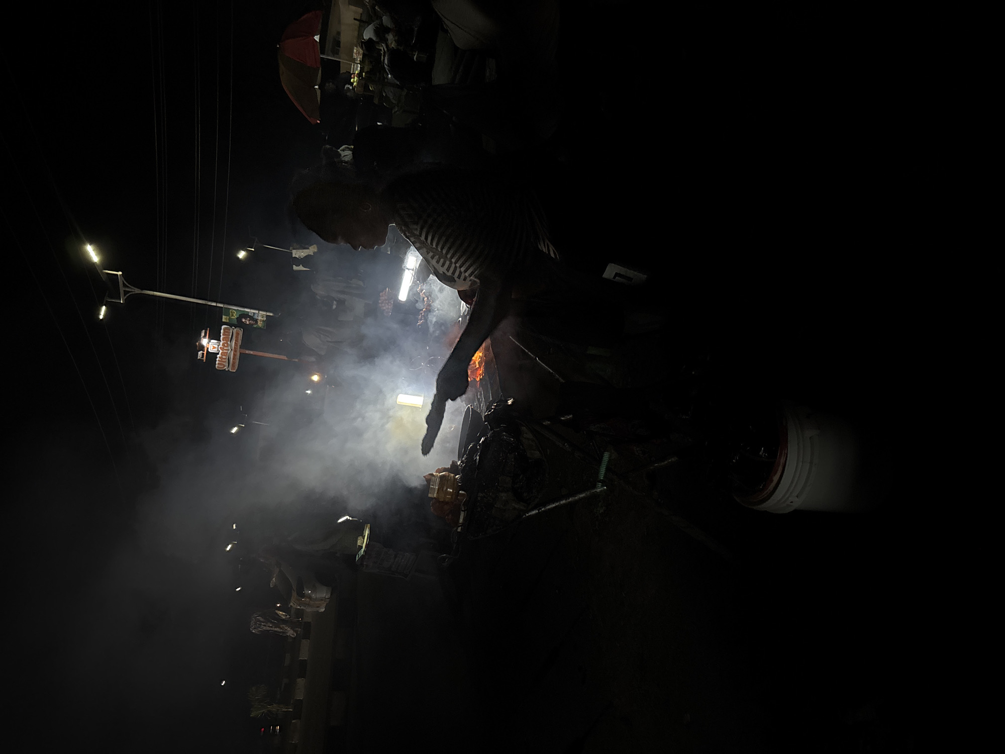 The image shows a street scene at night in Barakin Lerawi, Nigeria. A person wearing a striped shirt is bent over a grill, seemingly cooking food. There is a significant amount of smoke emanating from the grill, illuminated by the fire. Overhead electrical wires are visible, and a street sign for "Mr. Biggs" is seen. Streetlights are also present. There's an umbrella partially visible at the top of the frame and a white bucket to the right of the frame. The overall lighting is dim, with the fire providing the primary source of illumination.