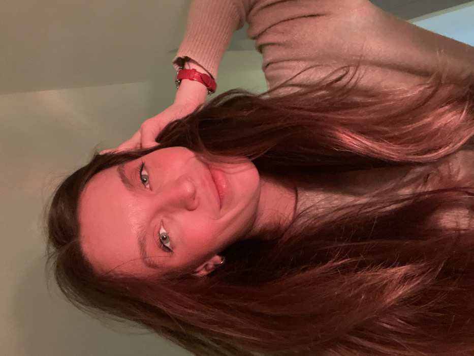 A woman with long, auburn hair smiles at the camera. She has light eyes, fair skin, and rosy cheeks. Her hand rests on her head, wrist adorned with a red watch. She wears a tan-colored sweater. The background appears to be a plain, light green wall. The image is slightly tilted, giving a casual, candid feel.