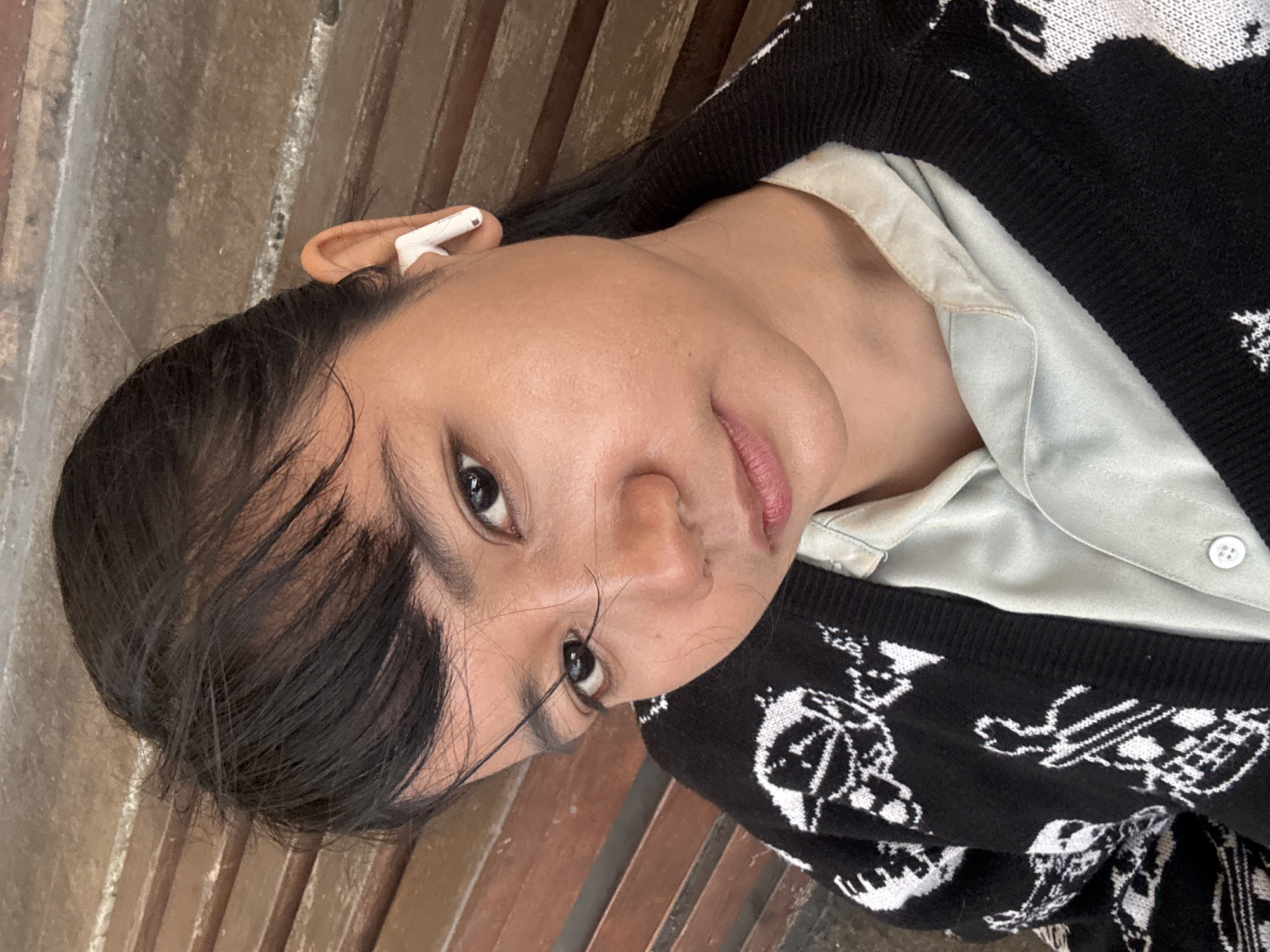 The image shows a close-up, slightly angled selfie of a young woman with dark hair, wearing a light-colored collared shirt under a black and white patterned cardigan. She has a white wireless earbud in her right ear. The background consists of wooden slatted seating, suggestive of a public waiting area or bench. The lighting appears to be natural, possibly from an overhead source. There is no visible indication of the specific location within North Jakarta, Indonesia.