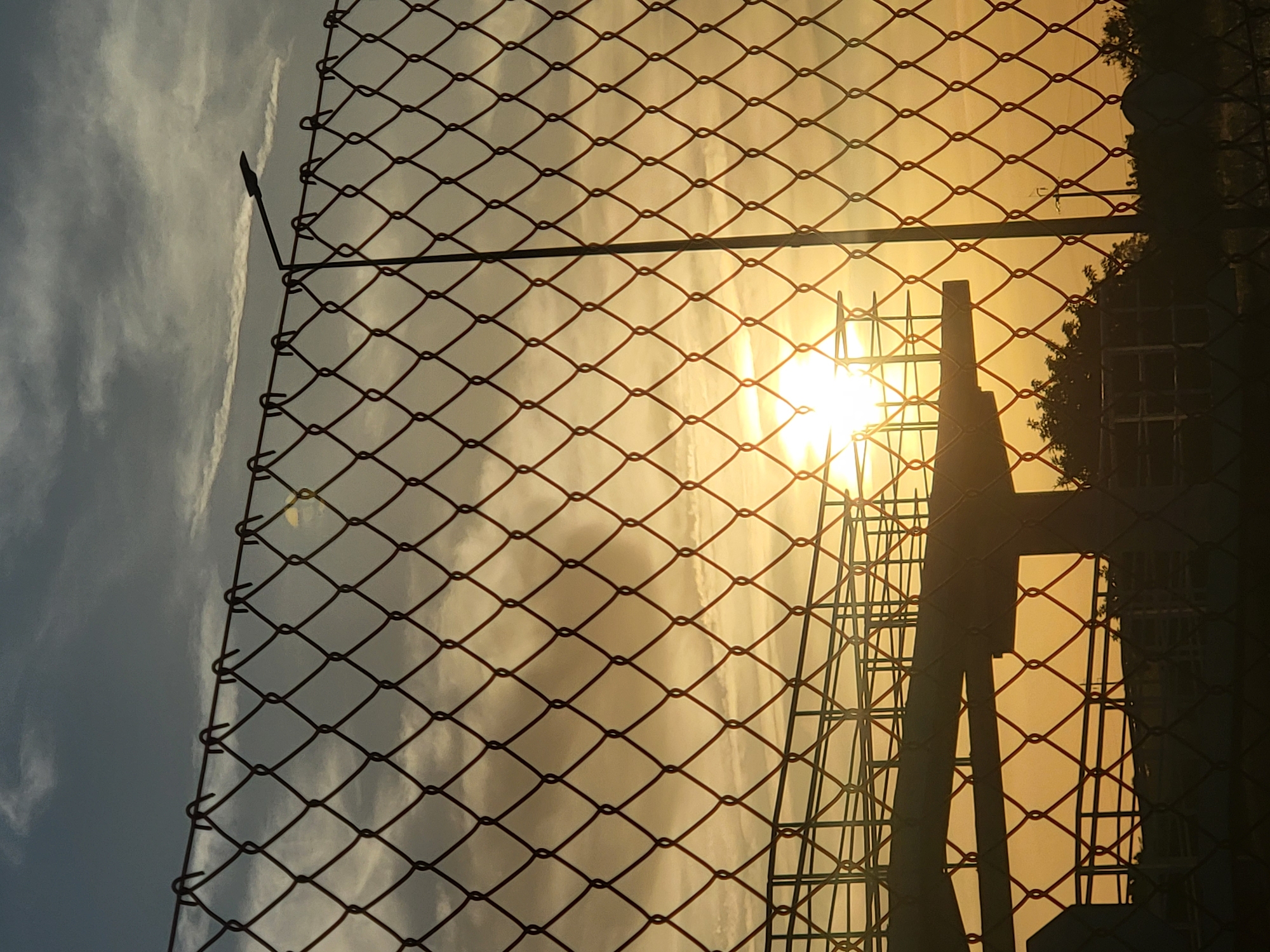 The image depicts a chain-link fence in the foreground, with a sunset visible through the diamond-shaped openings. In the middle ground, a silhouetted structure, possibly a bridge or overpass, is partially visible against the bright sky. The sun is a prominent feature, low on the horizon and casting a warm glow over the scene. The sky is a mix of blue and grey clouds, with streaks of white indicating atmospheric conditions. Trees are also visible in silhouette along the horizon line. A street light is mounted on a pole behind the fence, to the right of the structure. The overall impression is one of a partly industrial or urban landscape at dusk. The location is identified as Faro, Portugal.
