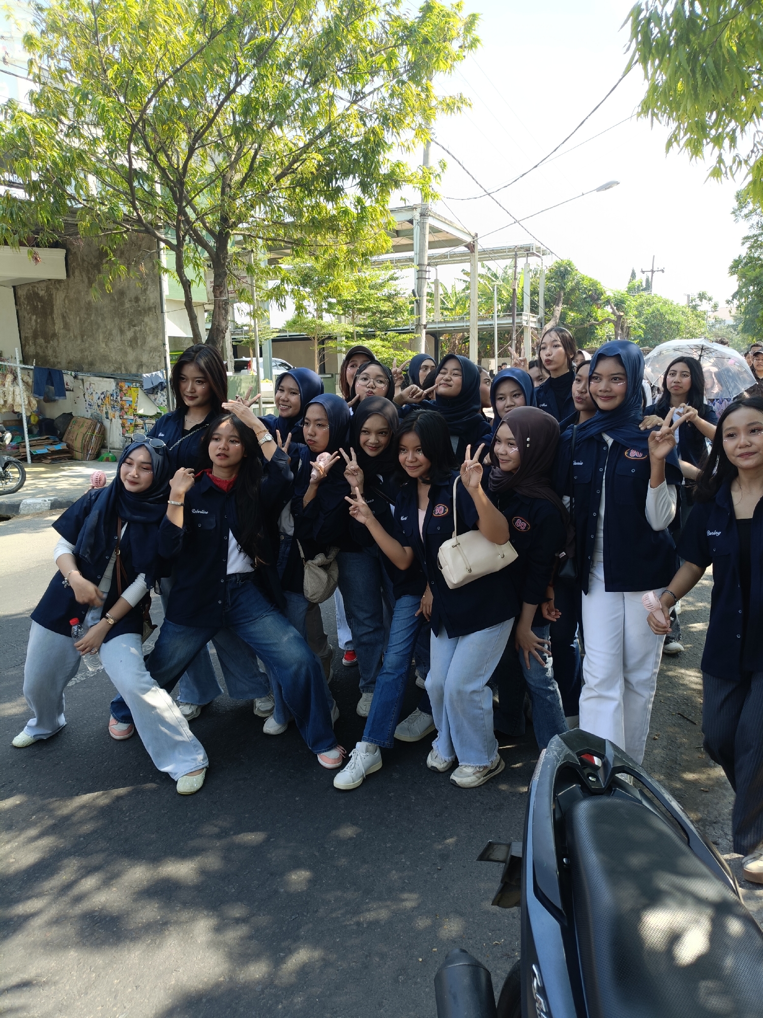 A group of young women, likely students, are posing for a photo outdoors in what appears to be a city street in Kediri City, Indonesia. They are mostly wearing dark blue collared shirts with an emblem on the left chest pocket, and denim jeans. Many are wearing headscarves, predominantly navy blue. Several women are making "peace" signs with their fingers, and some have glitter applied to their faces. One woman in the front center is wearing a red scarf. The background shows a building with some laundry hanging outside, trees with green foliage, a street light, and overhead electrical wires. The ground is asphalt, with some shadows from trees cast upon it. A black motorcycle is partially visible in the foreground, on the right side of the image. The lighting suggests it is daytime, likely morning or afternoon.