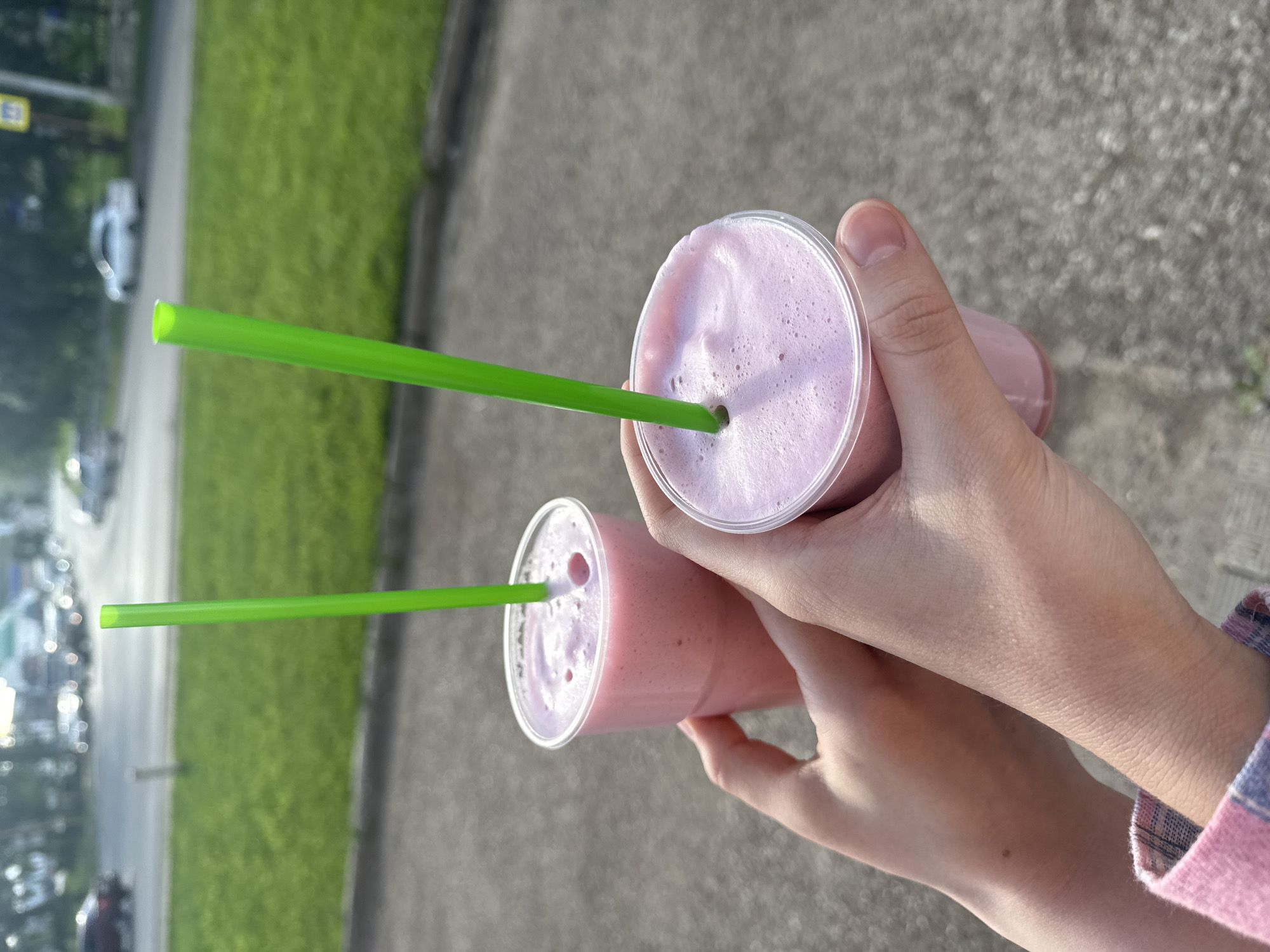 Two plastic cups of pink smoothie, each with a green straw, are held by two pairs of hands in the foreground. The cups are clear, and the smoothies are frothy. One cup is closer to the camera, and the other is slightly behind and to the right. The background is blurred and shows a street with cars and green grass. The ground in the foreground is a gray, concrete surface. The scene appears to be outdoors in an urban environment during the daytime.