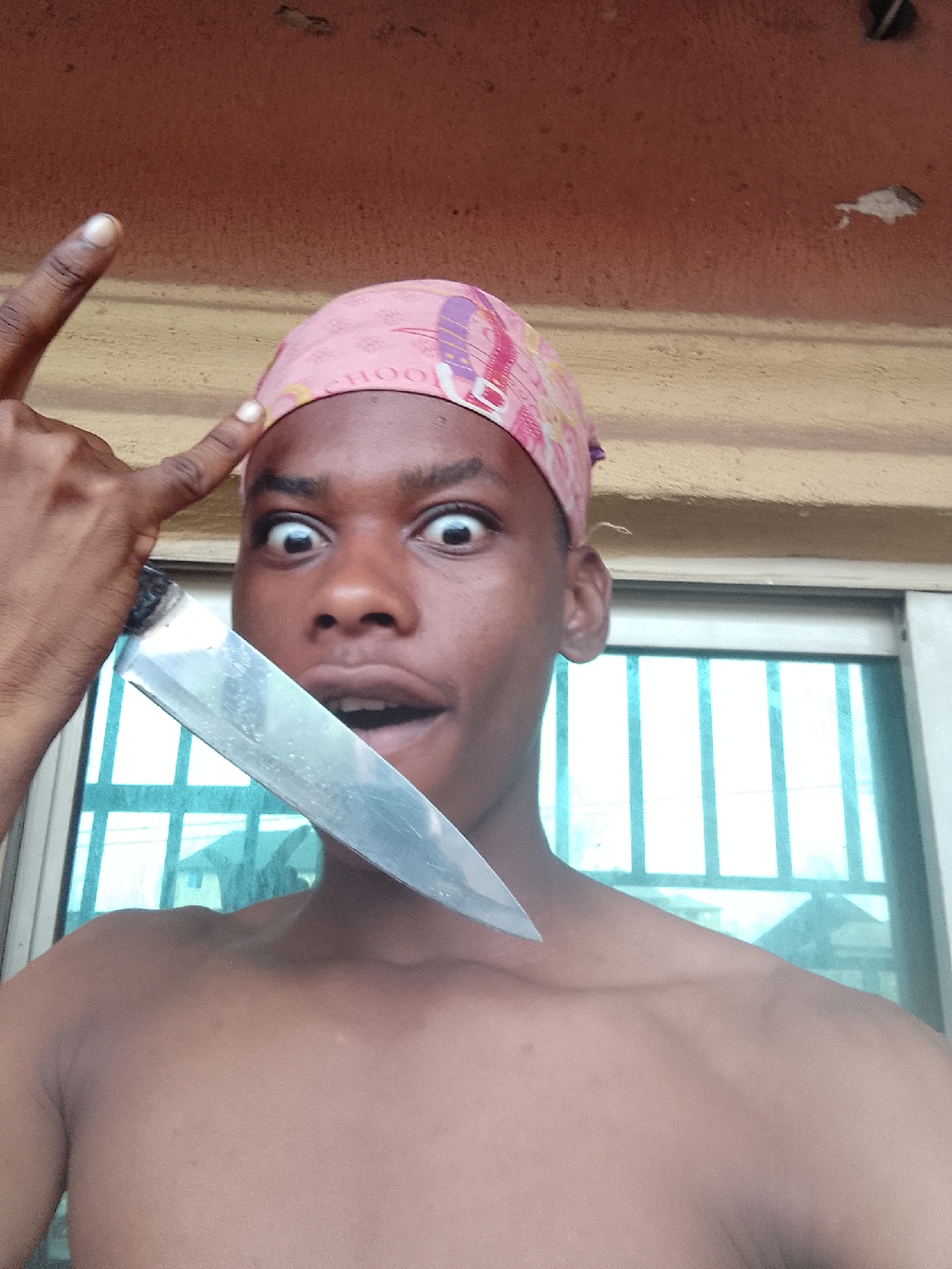 A shirtless dark-skinned male with a pink bandana on his head is holding a large kitchen knife near his face, with the blade pointing towards his neck. He is making a surprised expression with wide eyes and an open mouth. His left index finger is pointed towards his head. The background includes a wall with a window. The location is Nkpor, Nigeria.