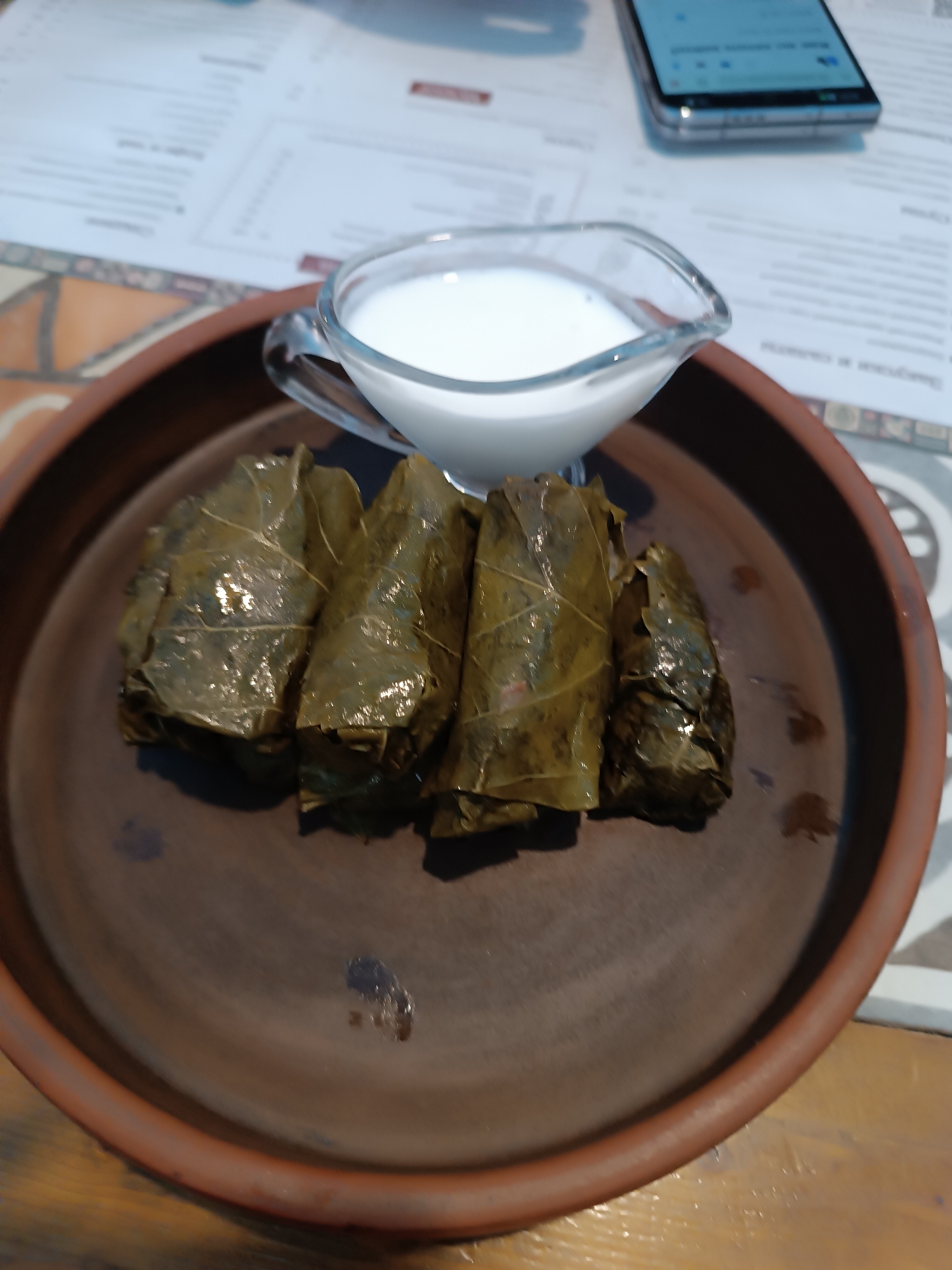 A plate of dolma with a side of yogurt sauce is on a table in a cafe. There are five dolma rolls wrapped in grape leaves arranged in a row on a brown ceramic plate. A small glass gravy boat filled with white yogurt sauce rests on the plate next to the dolma. In the background, a smartphone lies on top of a menu. The location is Izhevsk, Russia.