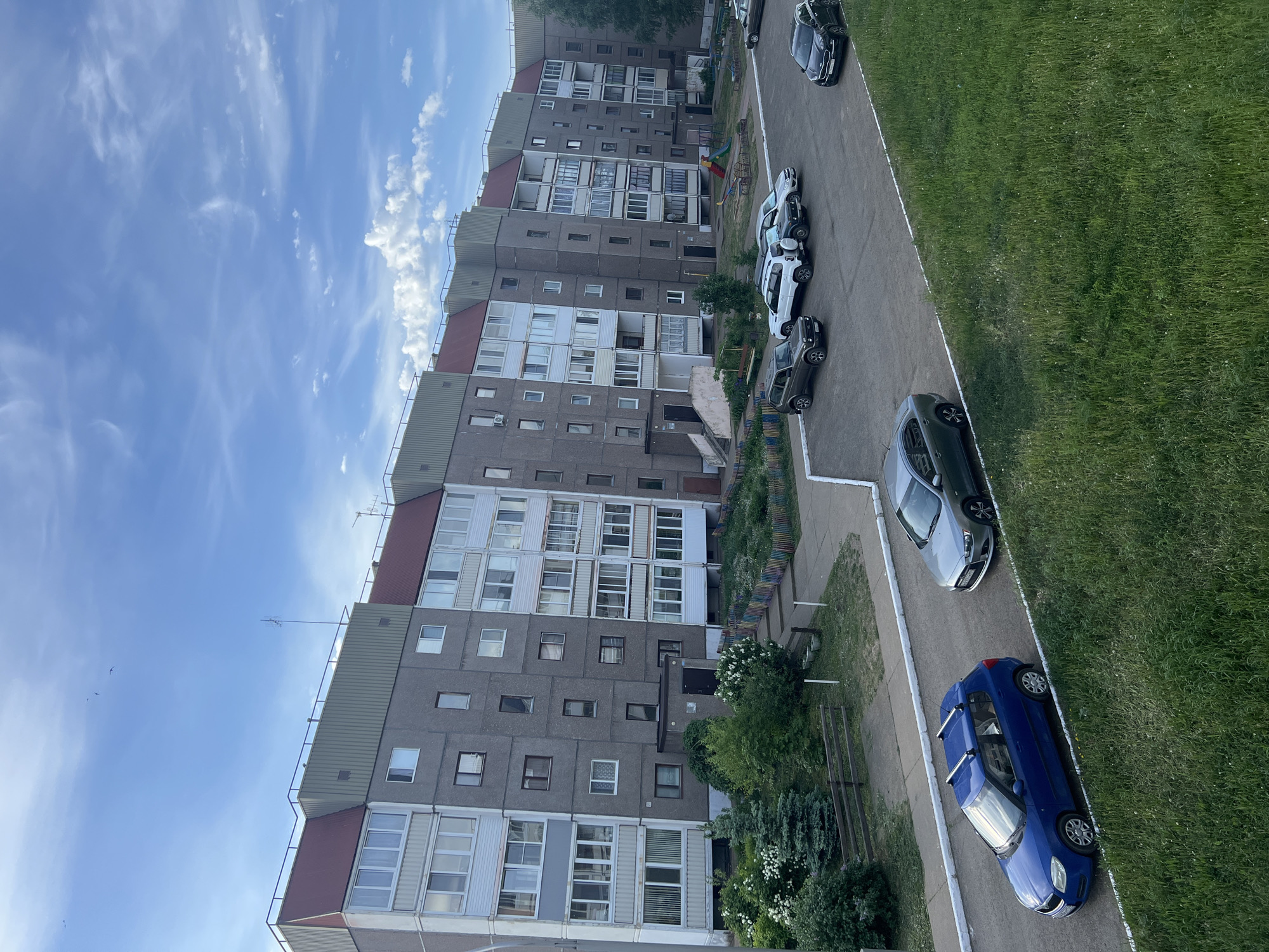 The image shows a row of Soviet-era apartment buildings under a blue, slightly cloudy sky, most likely located in the city of Марковский, Russia. The buildings are concrete, six to seven stories high, with a mix of grey and white facades. A paved road runs parallel to the buildings, with several parked cars including one blue and one silver car that are facing toward the camera. A strip of grass is visible on the right edge of the frame, bordering the road. There are trees and vegetation in the foreground and between the buildings. In the background, a playground is visible between the apartment blocks.