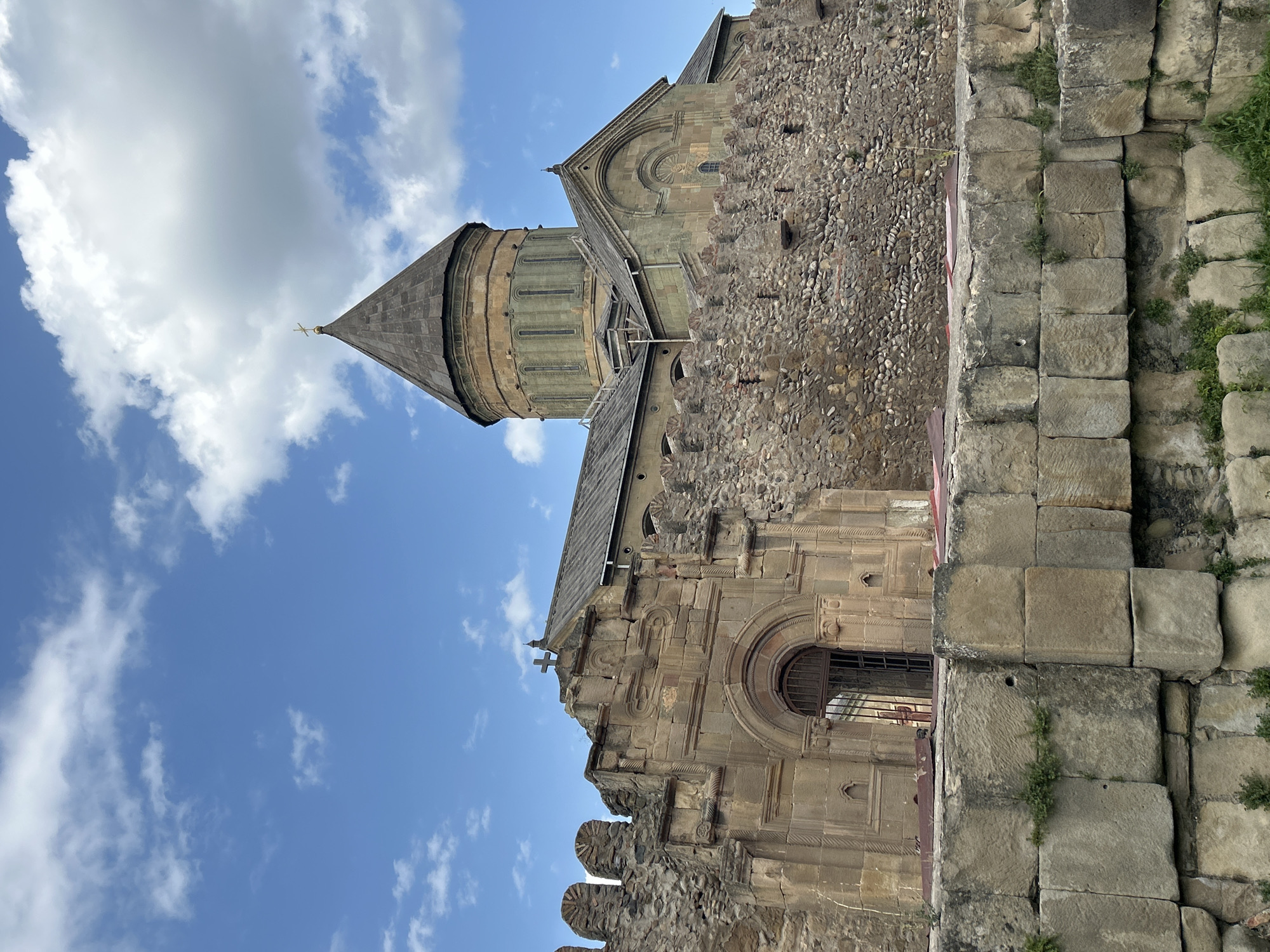 The image captures the Svetitskhoveli Cathedral, a prominent landmark in the city of Mtskheta, Georgia. The cathedral, constructed from stone, features a large, conical dome topped with a golden cross. The surrounding structure includes weathered stone walls and fortifications, some of which are crumbling. The sky is blue with scattered white clouds. The foreground is dominated by a low stone wall composed of large, roughly hewn blocks, with patches of green vegetation growing between the stones. The overall scene depicts an ancient religious site under a clear sky.
