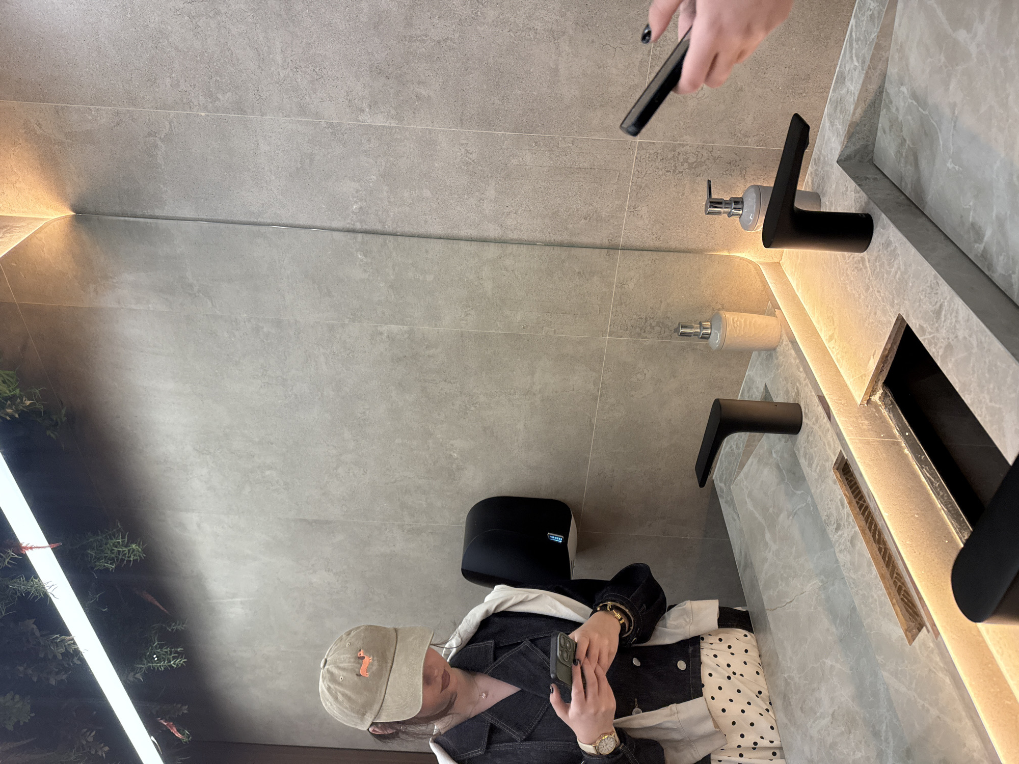 A person wearing a khaki-colored baseball cap with an orange fox logo, a denim jacket, and a polka-dotted skirt is holding a cell phone in a restroom. The restroom features a marble countertop with a modern black faucet and a soap dispenser. The restroom also has a black paper towel dispenser mounted on the wall. The reflection of a person holding a cell phone is visible in the mirror above the countertop. There is an illuminated wall with plants behind the person in the reflection. The location is Yerevan, Armenia.