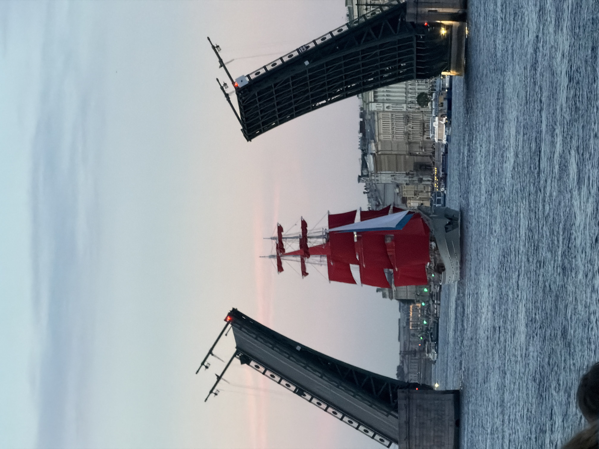 In Saint Petersburg, Russia, at dusk, a large sailing ship with red sails passes through an opened drawbridge. The bridge is raised in two sections, forming an angle above the water. The ship, a three-masted schooner, is painted white with red sails and a white and blue flag. Buildings line the distant shore. Several smaller boats are visible on the water. The sky is a soft gradient of pale blue and pink. The water is rippled.