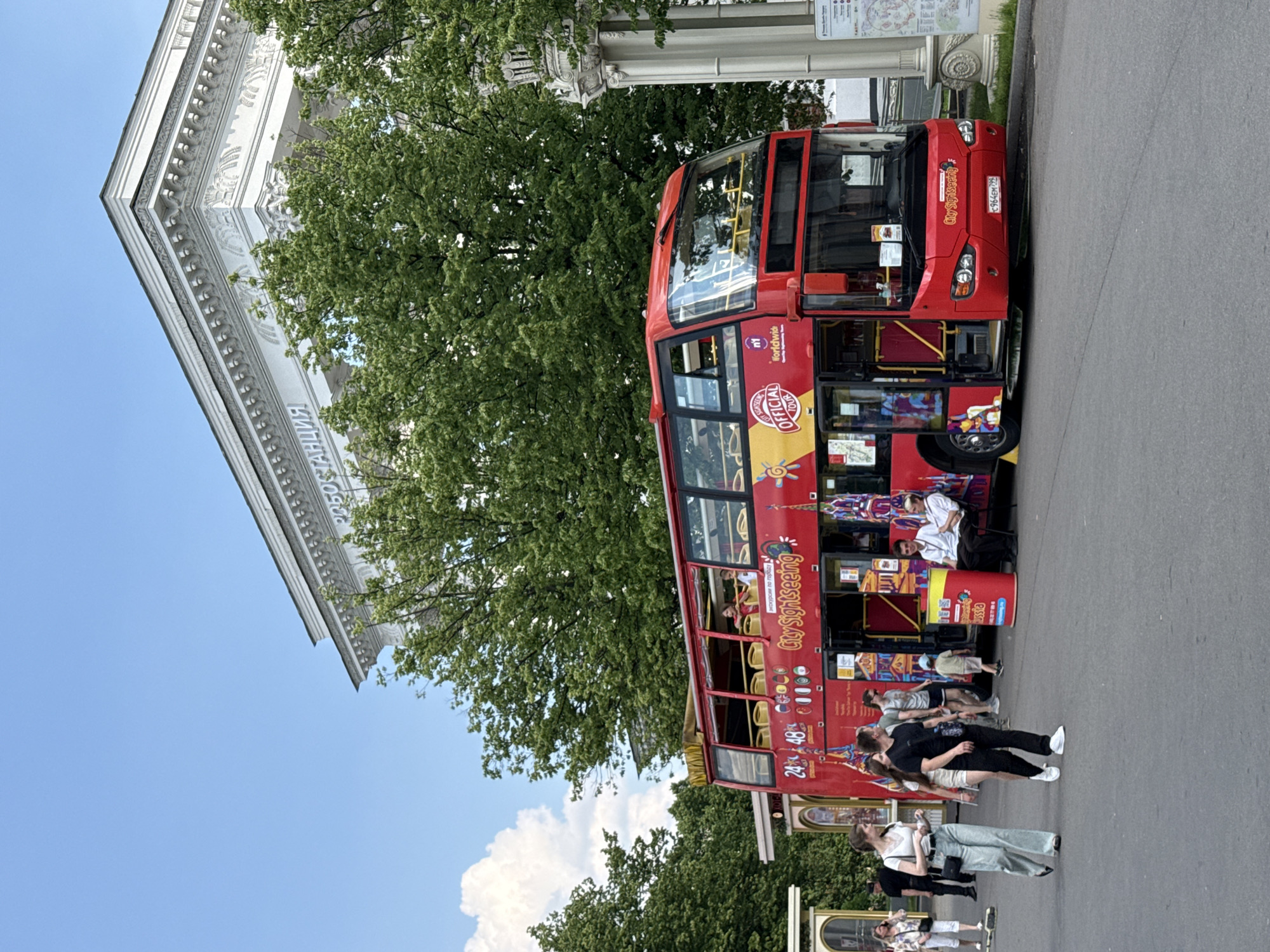 The image depicts a red double-decker tour bus parked on an asphalt road. The bus has "City Sightseeing" branding and "OFFICIAL TOUR" logos. Several people are visible, some walking on the sidewalk and others on the bus. A large, ornate white building with Russian text, which appears to be "РОБОСТАНЦИЯ" (Robostantsiya), is in the background, partially obscured by lush green trees. The sky is clear and blue. The scene suggests a tourist area in Moscow, Russia, likely near an exhibition center or a significant historical site, given the architecture and the presence of a tour bus.