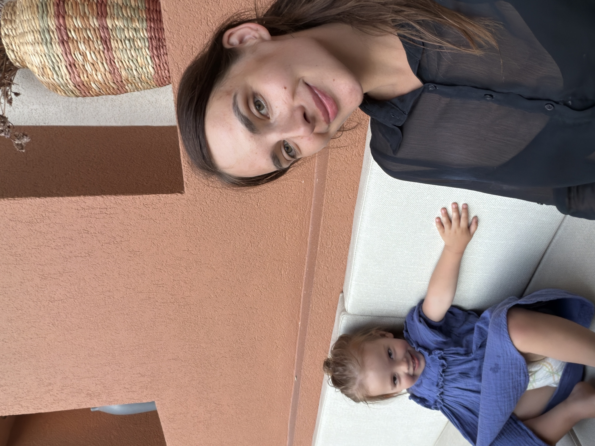 The media file is an image of two subjects, a woman and a young child, on a couch outdoors. The woman is in the foreground, looking towards the camera with a slight smile. She has long, brown hair and is wearing a sheer black blouse. Next to her, lying on the couch, is a young child in a blue dress. The child is also smiling and looking at the camera. The background consists of a textured orange wall and a woven basket. The lighting suggests it is daytime. The overall mood is relaxed and intimate, possibly capturing a moment of family time. The location is Fethiye, Turkey.